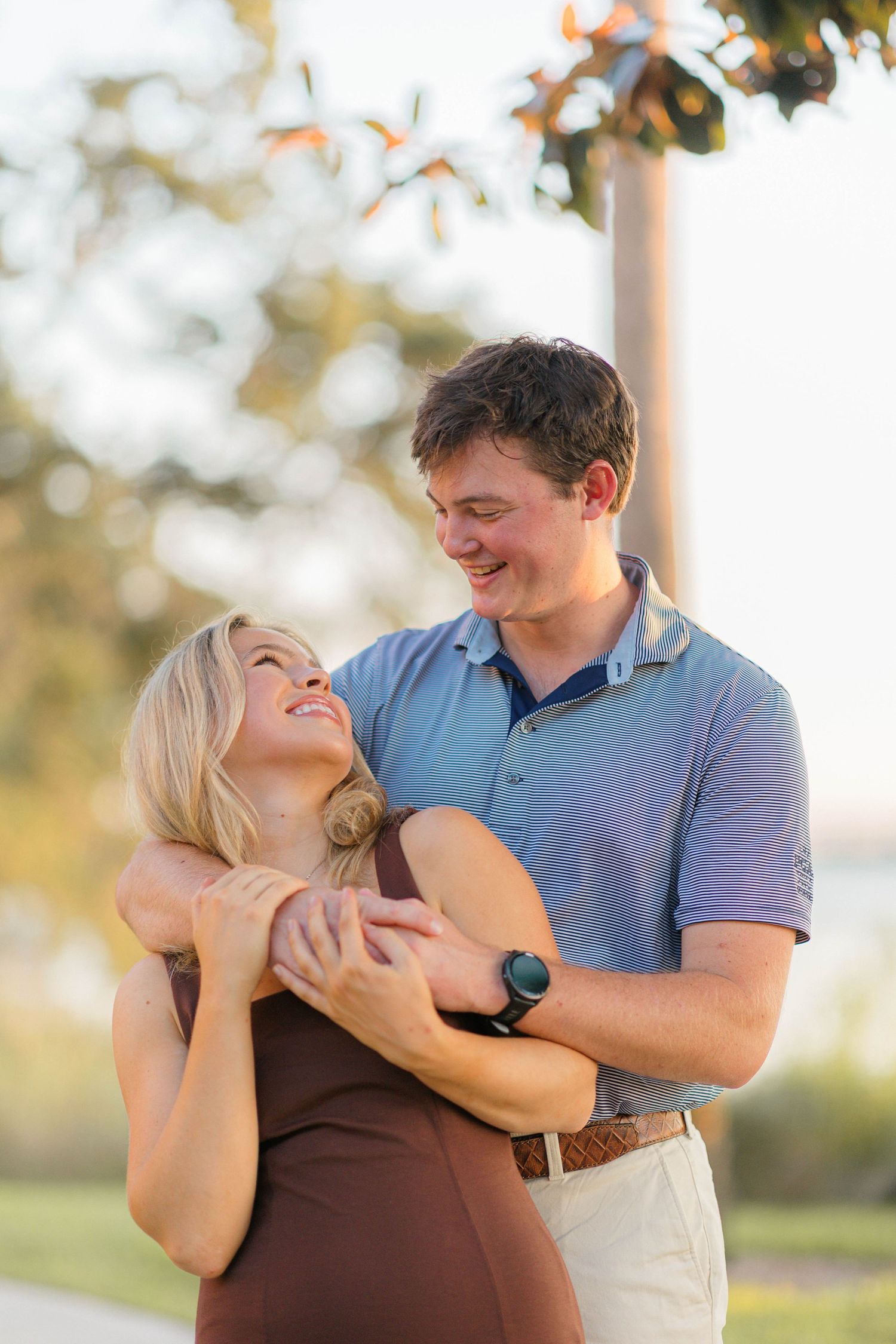 Happy couple embracing outdoors, woman in brown dress laughing as man in blue polo hugs her from behind.