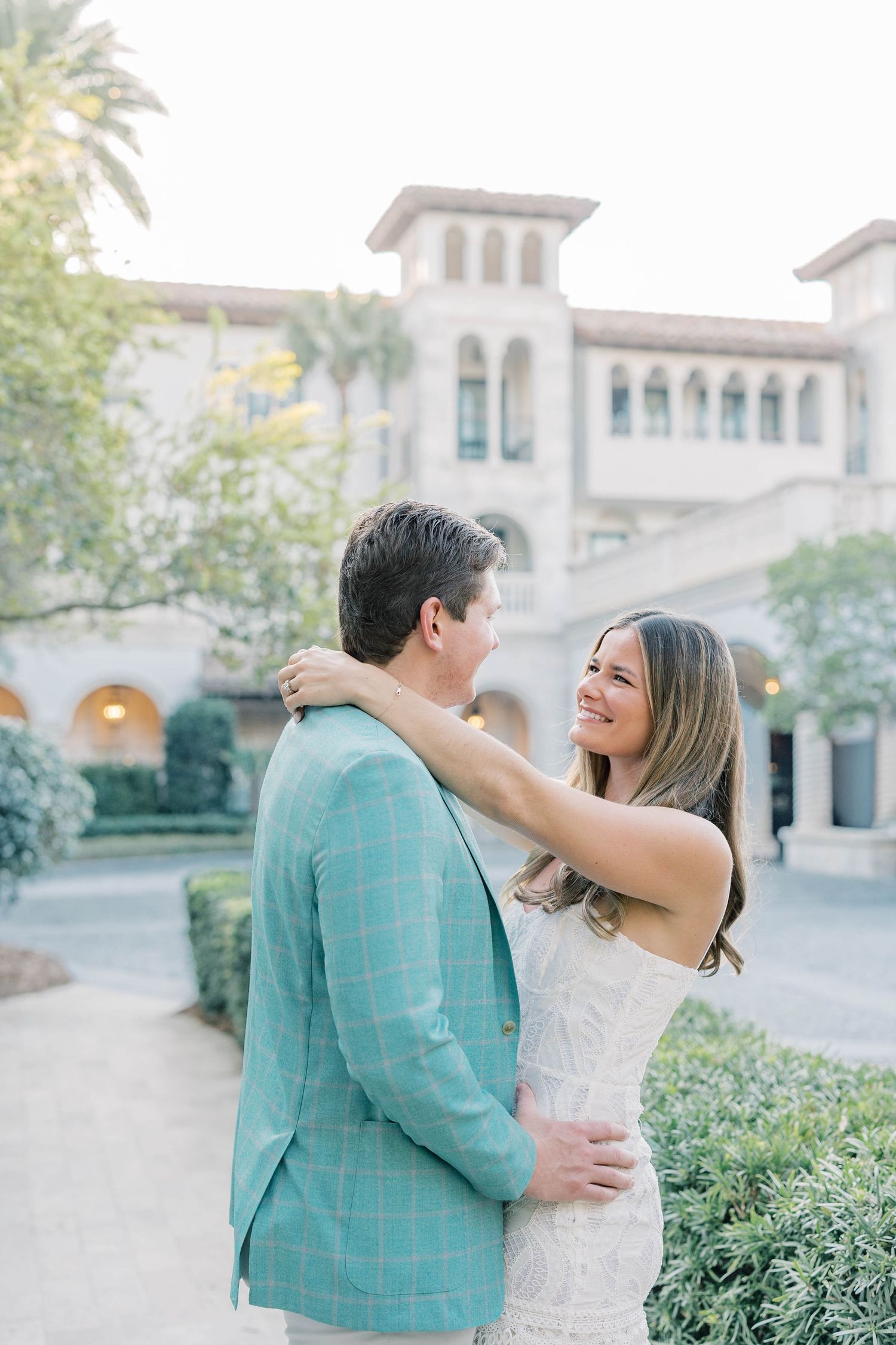 A smiling couple embraces outdoors near an elegant Mediterranean-style building, the woman in white dress, man in teal plaid blazer.