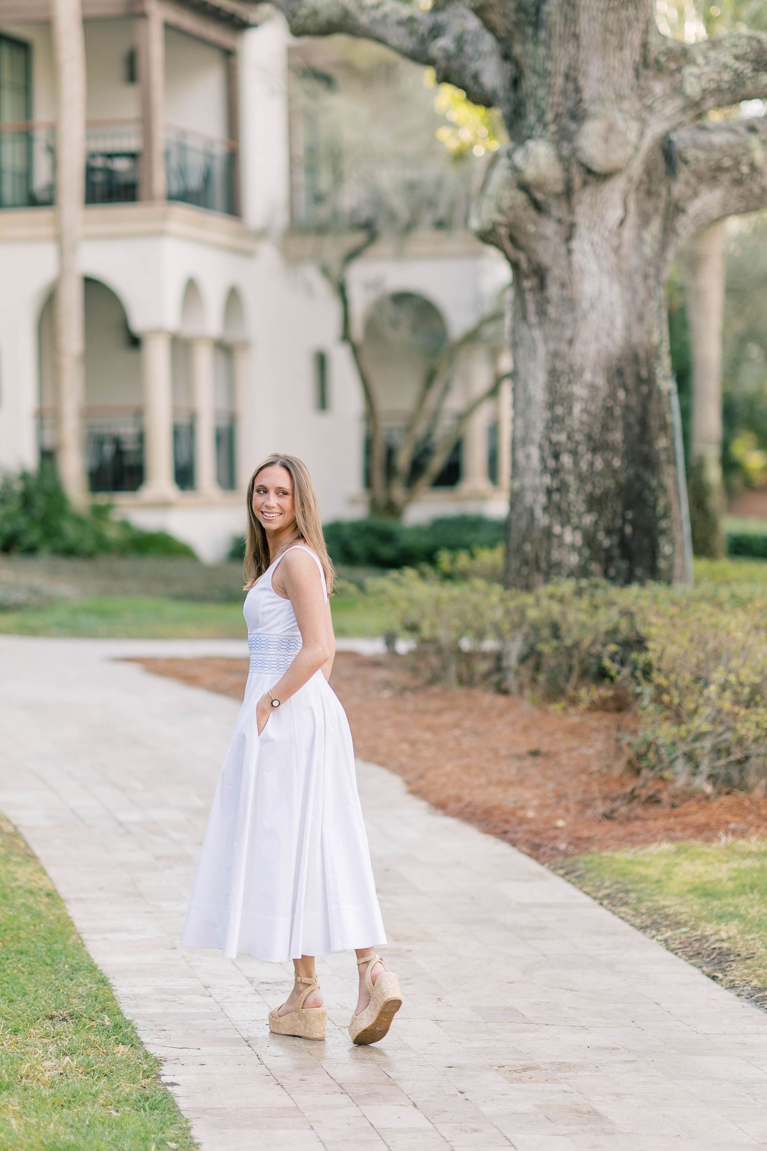 White sundress flowing in breeze along stone pathway with trees and architecture in background.