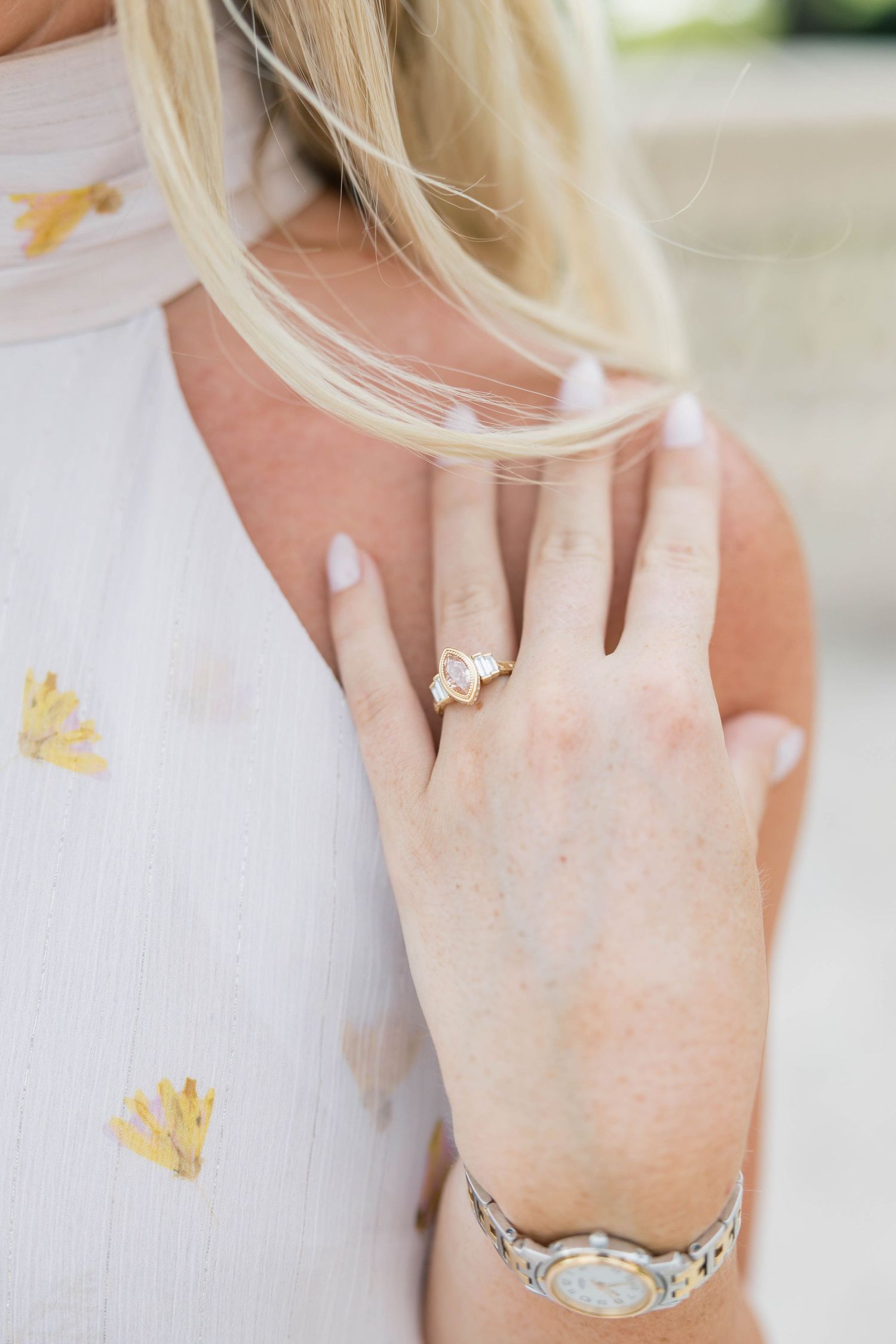 Close-up of a woman's hand wearing a gold ring and two-tone watch, with blonde hair and white floral top.