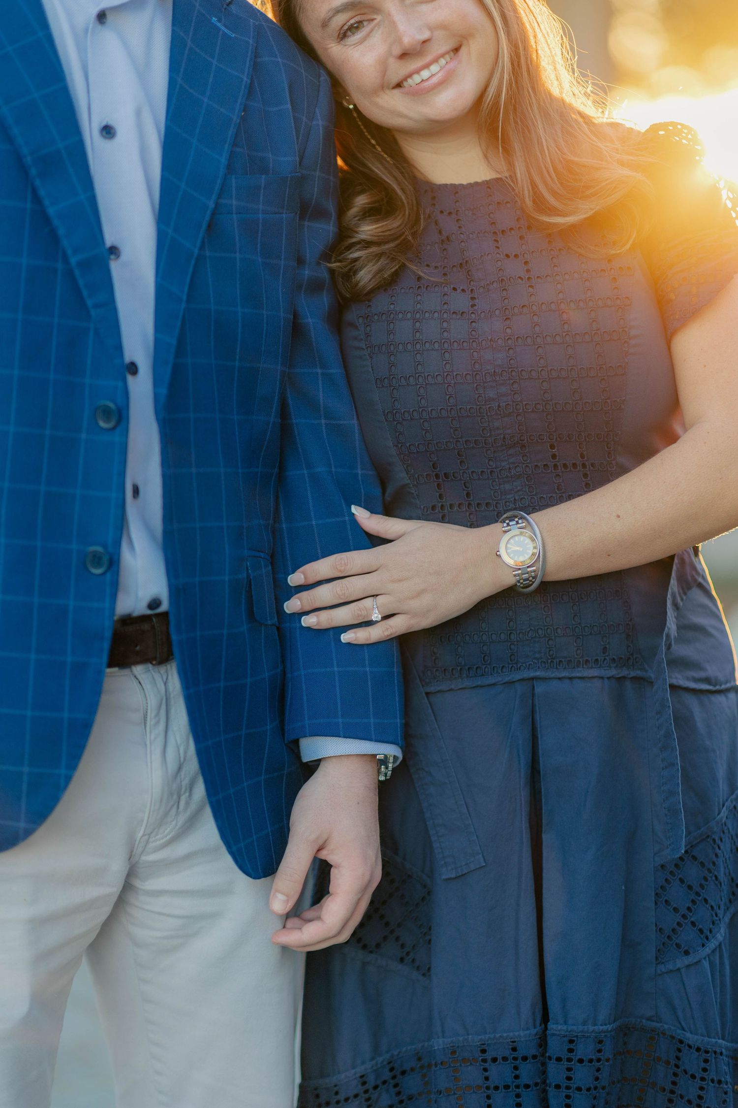 Couple in coordinating blue outfits posing at golden hour, woman showing engagement ring and silver watch.