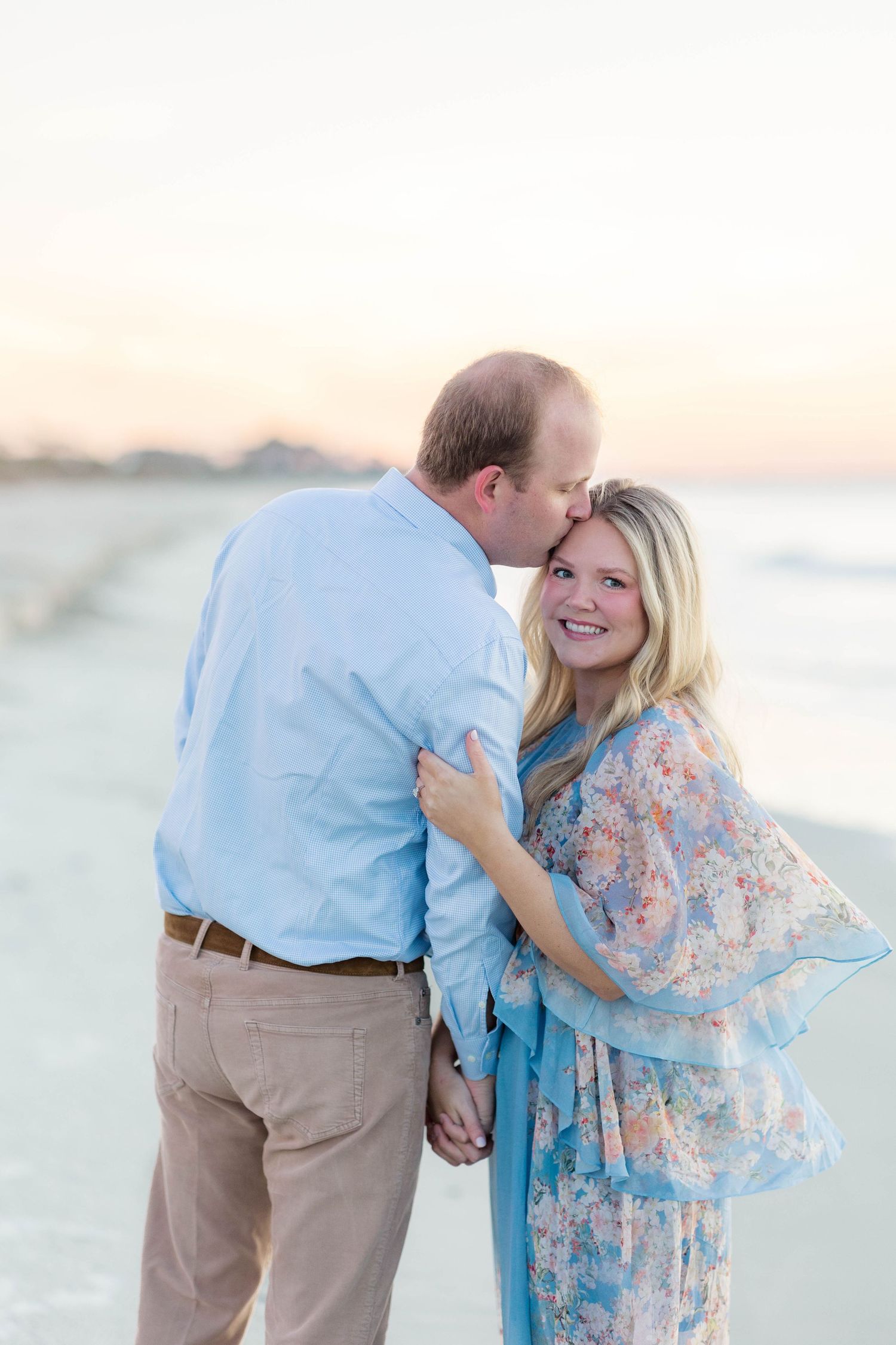 Couple sharing a romantic kiss on the beach at sunset, woman in floral blue dress smiling, man in light blue shirt.