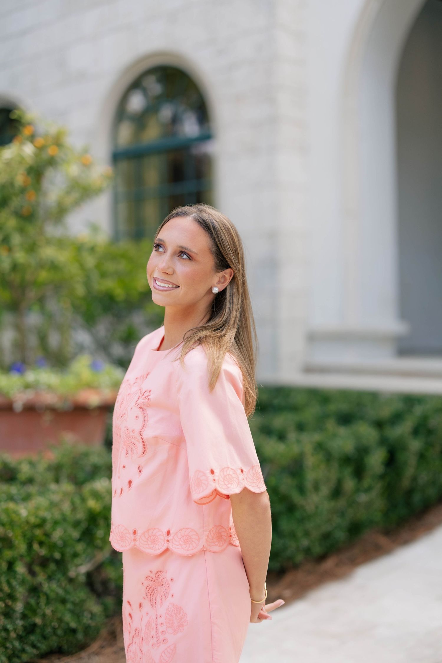 Person in coral pink lace top posing outdoors near white building with arched windows.