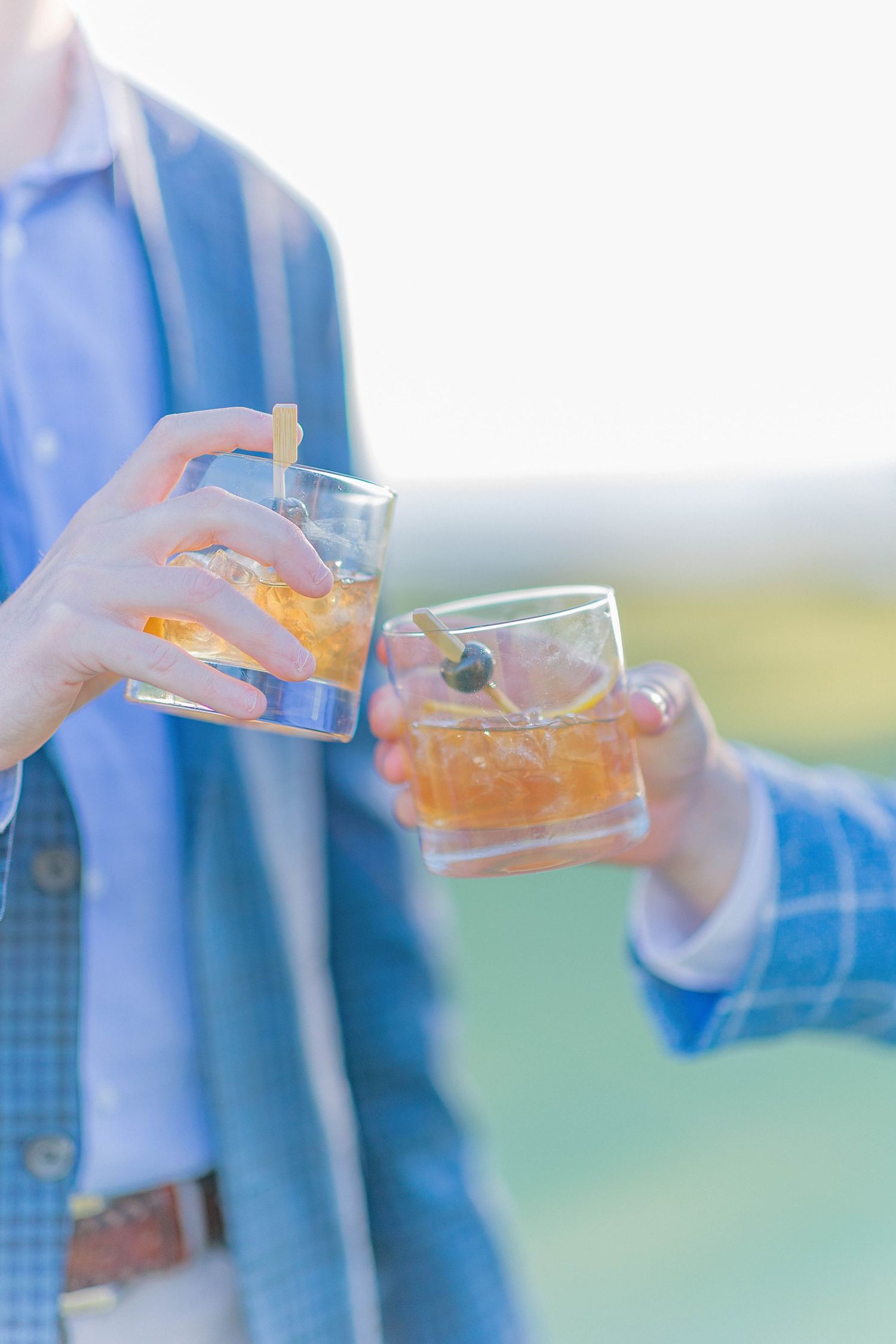 Two hands holding whiskey cocktails with ice outdoors, wearing blue blazers.