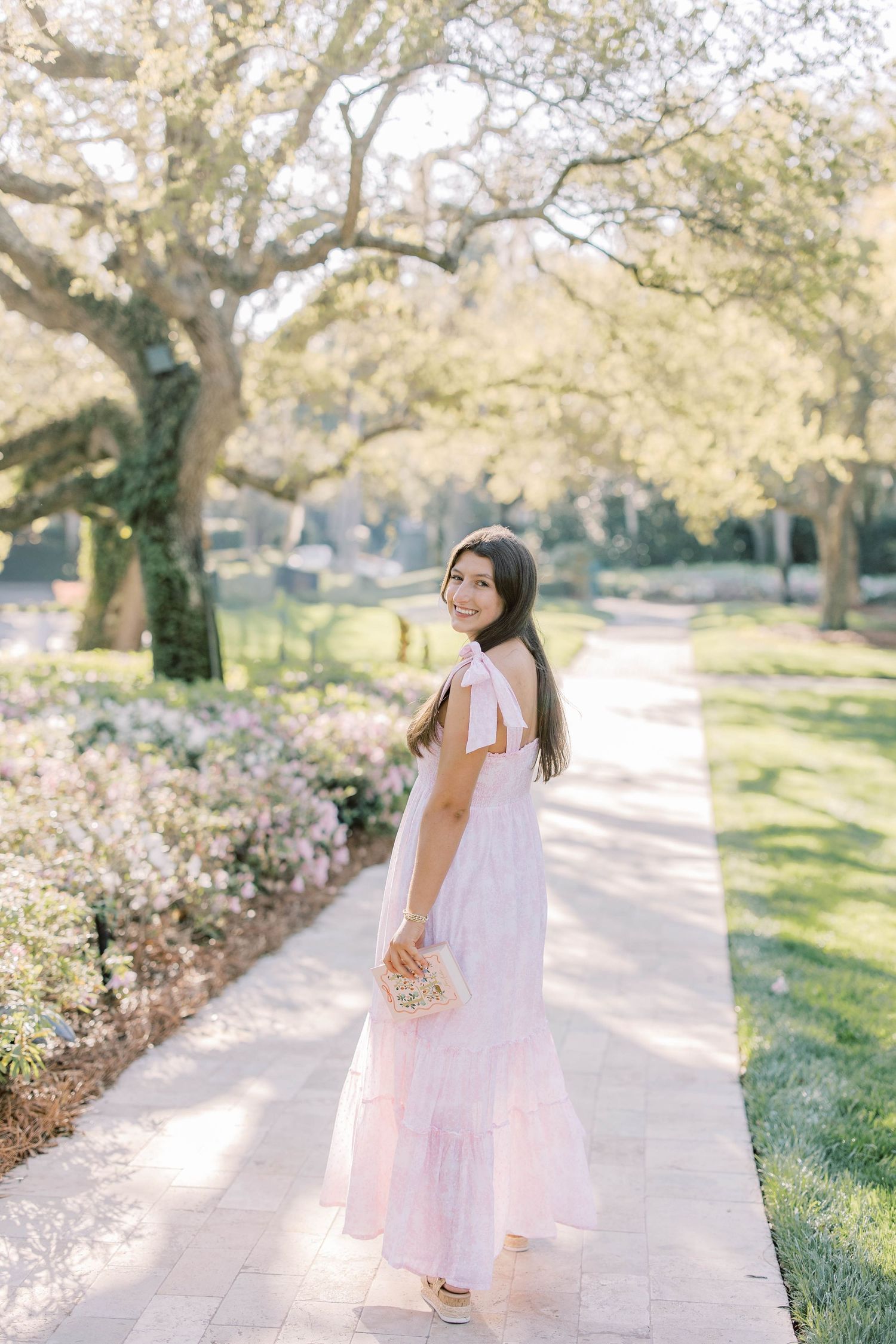 Young woman in pink maxi dress smiling on a sunlit garden path surrounded by blooming flowers and trees.