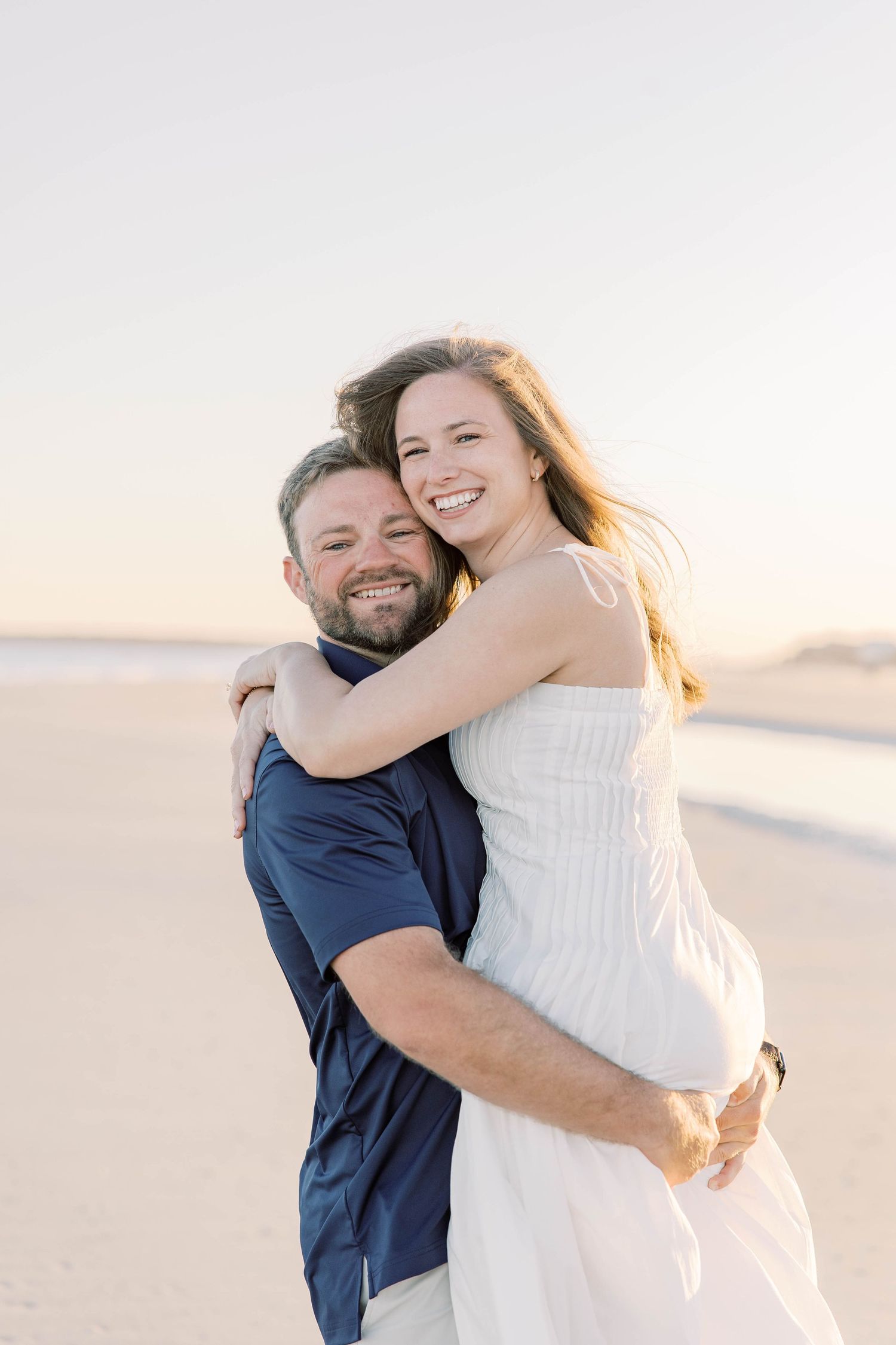 Happy couple embracing on a sandy beach at sunset, woman in white dress and man in navy shirt smiling.