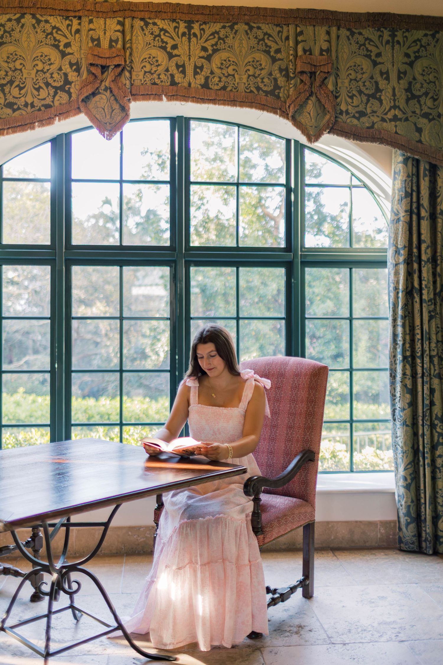 Woman in pink dress reading a book at a table by an ornate arched window.