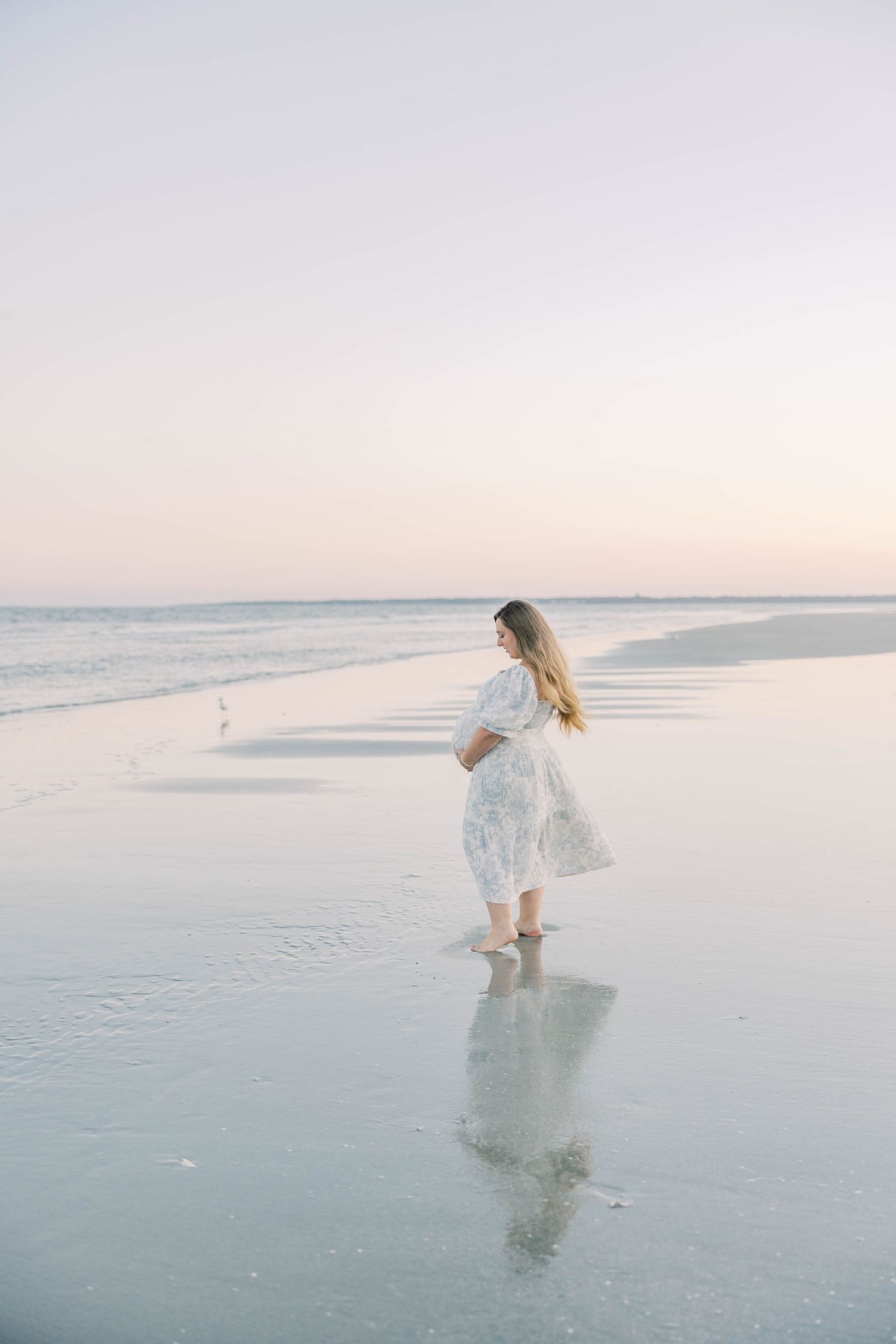 Pregnant woman in white lace dress standing barefoot on a serene beach at sunset with soft pastel sky.