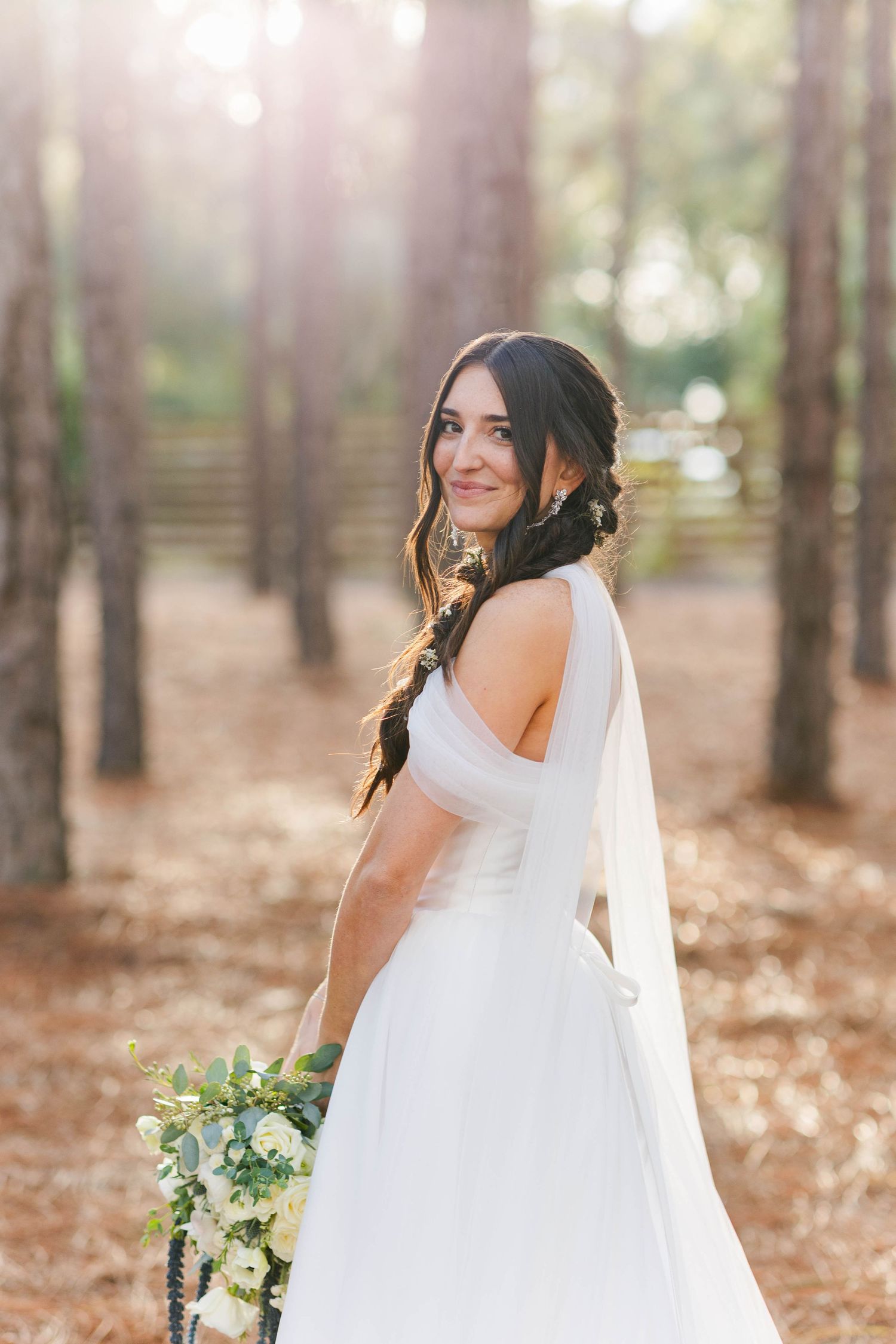 Bride in flowing white wedding dress poses elegantly in a sunlit pine forest holding a white and green bouquet.