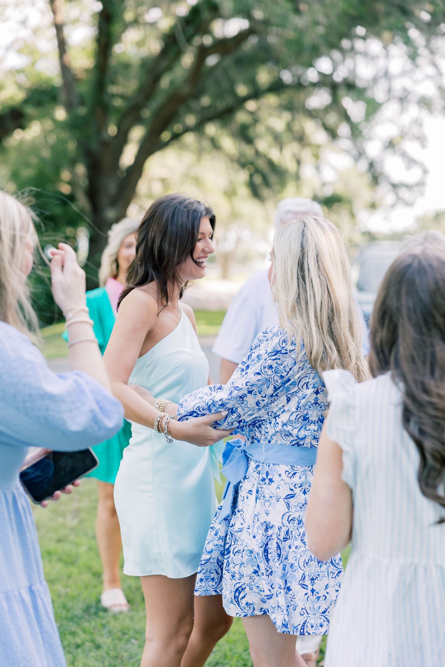 Women in summer dresses socializing outdoors at a garden party under shaded trees on a sunny day.
