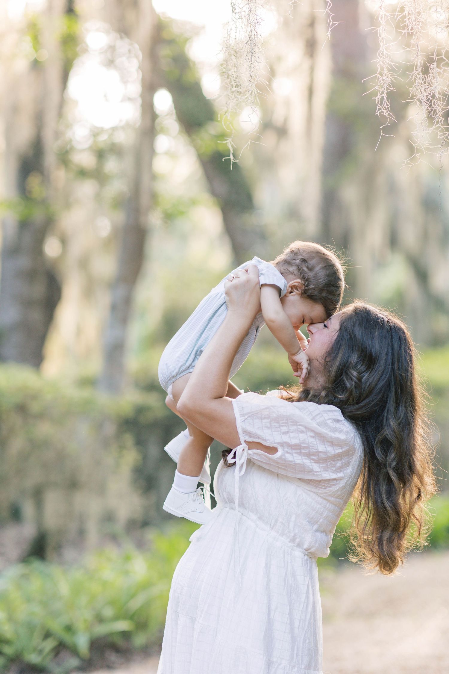 A joyful mother in a white dress lifts her baby outdoors among sunlit trees with Spanish moss.
