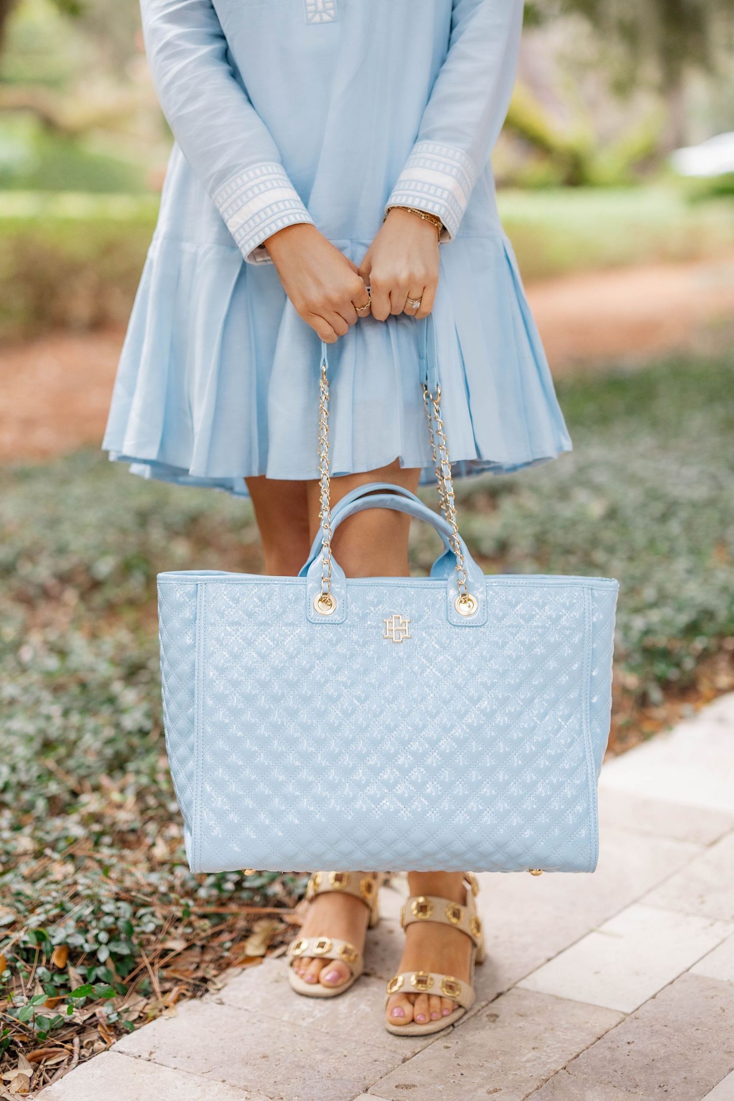 Woman in light blue dress holding a matching quilted blue tote bag with gold chain straps, wearing gold sandals.
