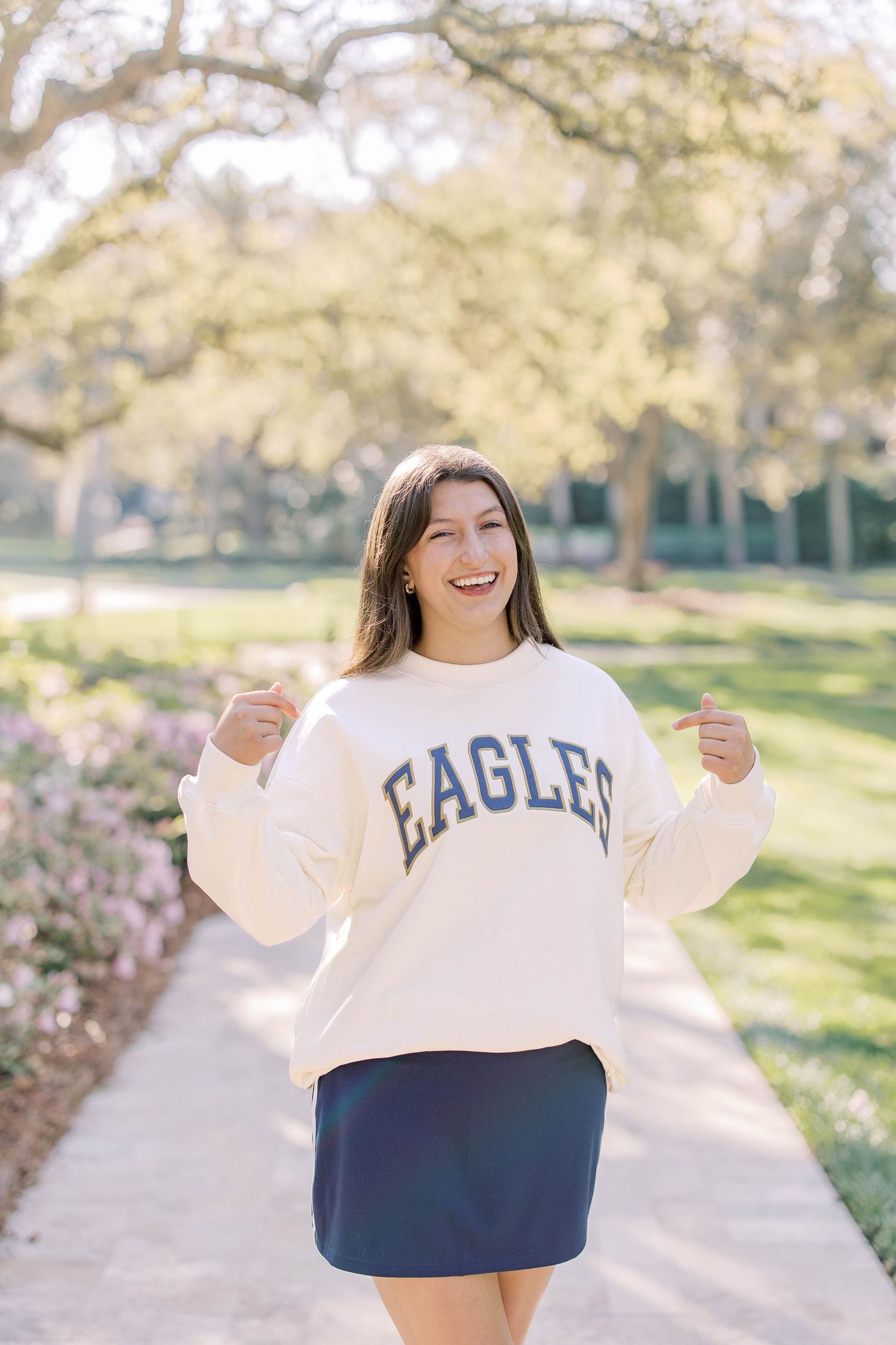 Smiling young woman in cream Eagles sweatshirt and navy skirt posing outdoors in a tree-lined park pathway.