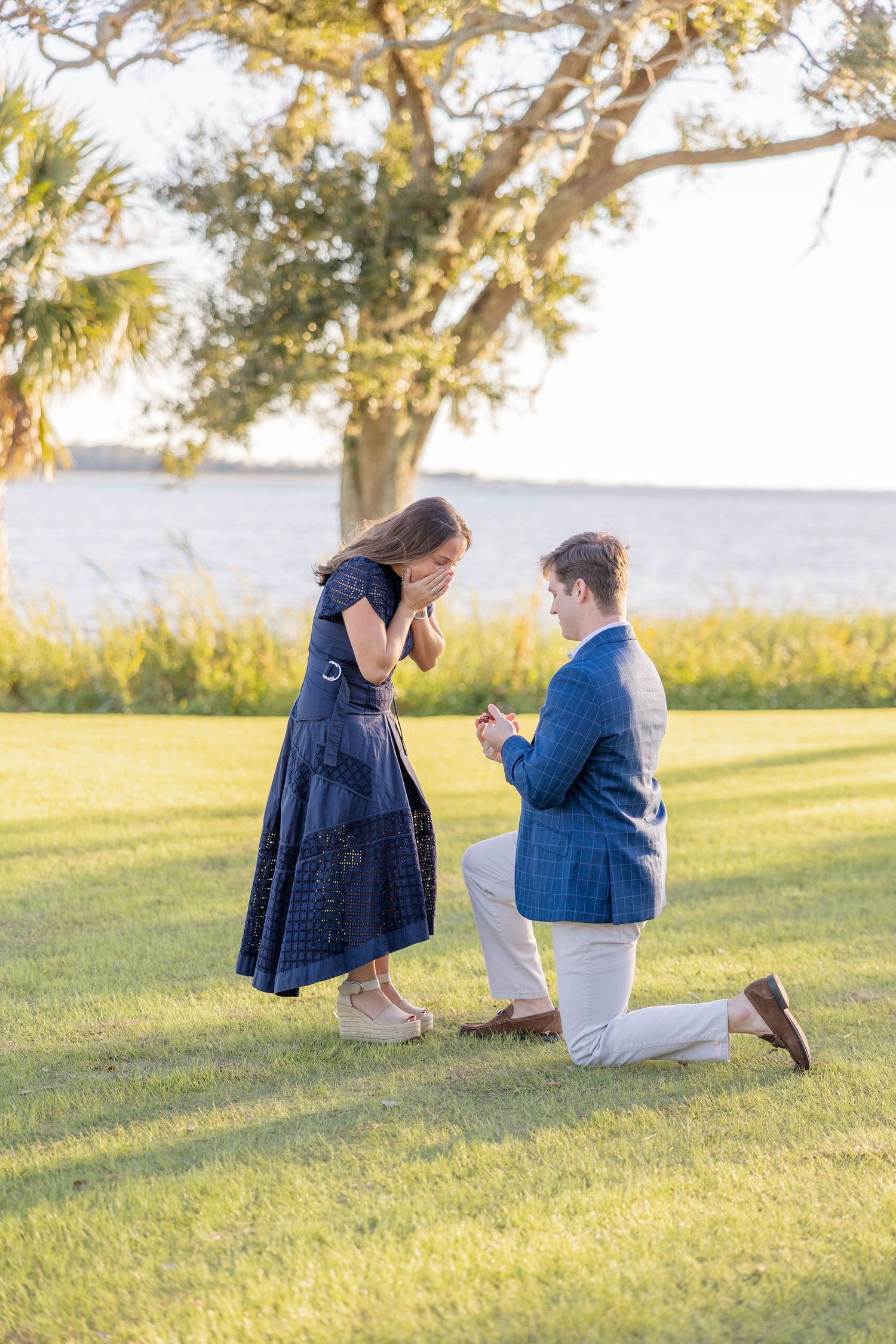 Man in blue blazer proposes on one knee to woman in navy dress outdoors near waterfront at golden hour.