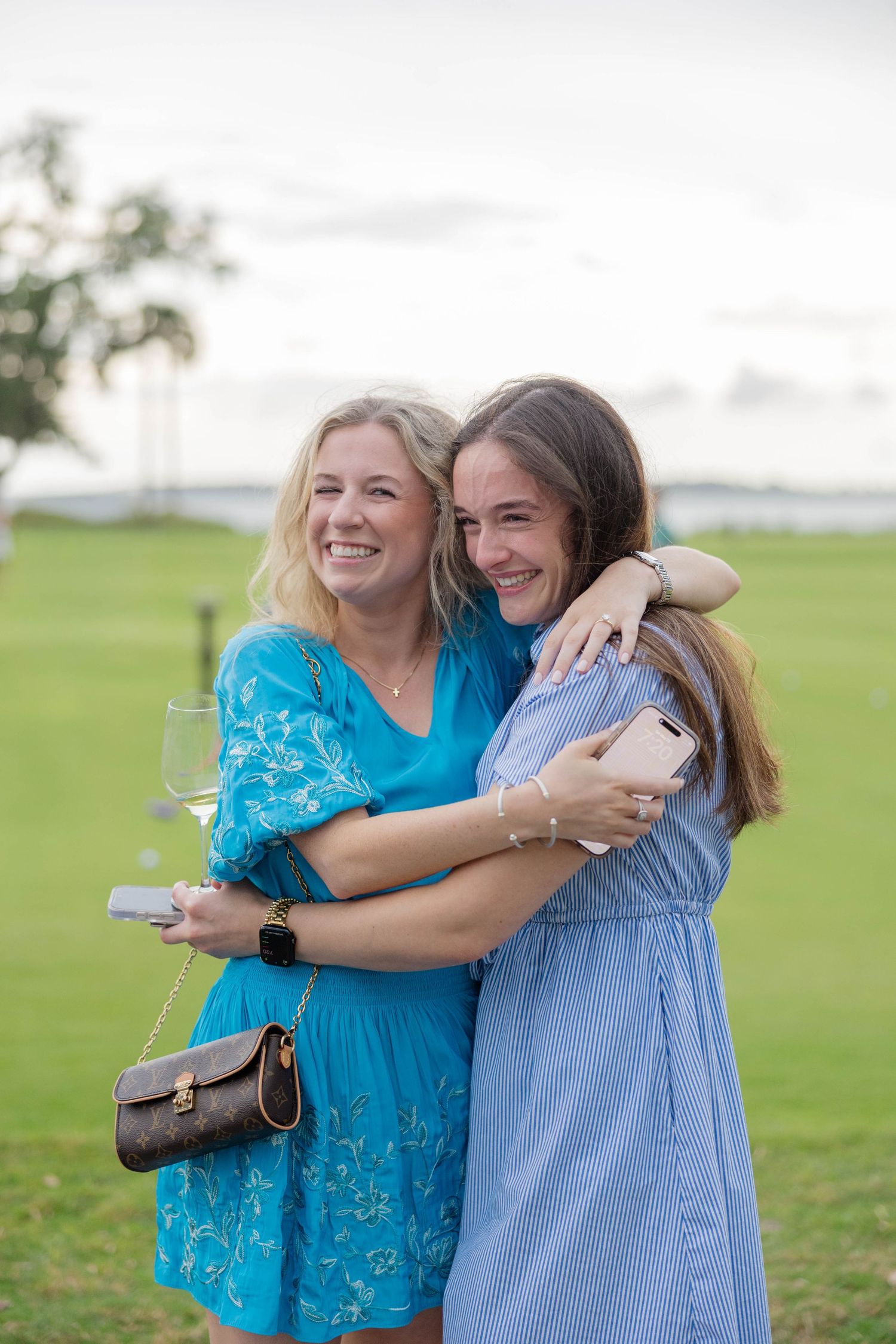 Two women in blue dresses hugging and smiling outdoors on a green lawn at dusk.