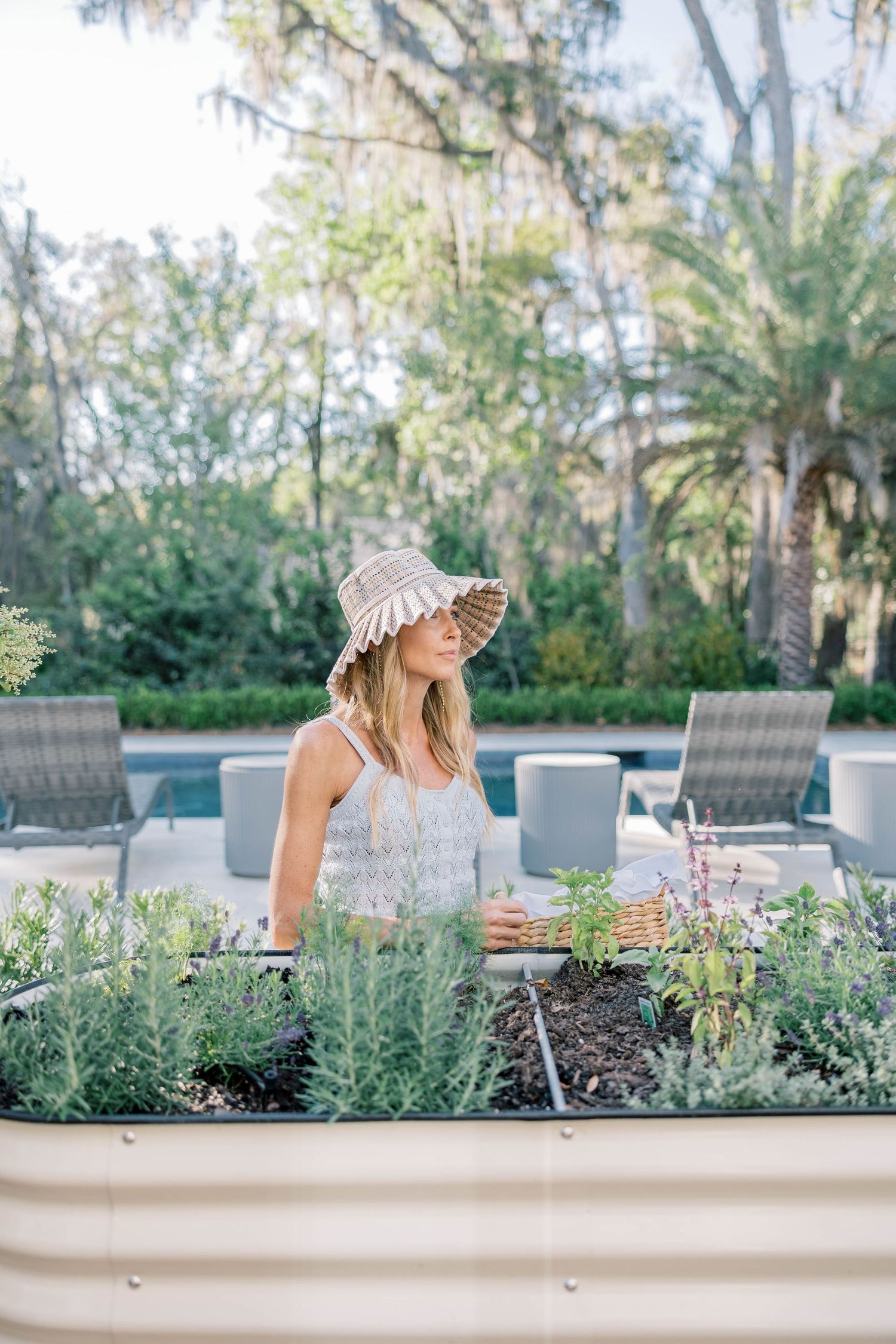 Woman in straw hat tending raised garden bed with herbs near pool, surrounded by lush greenery and palm trees.