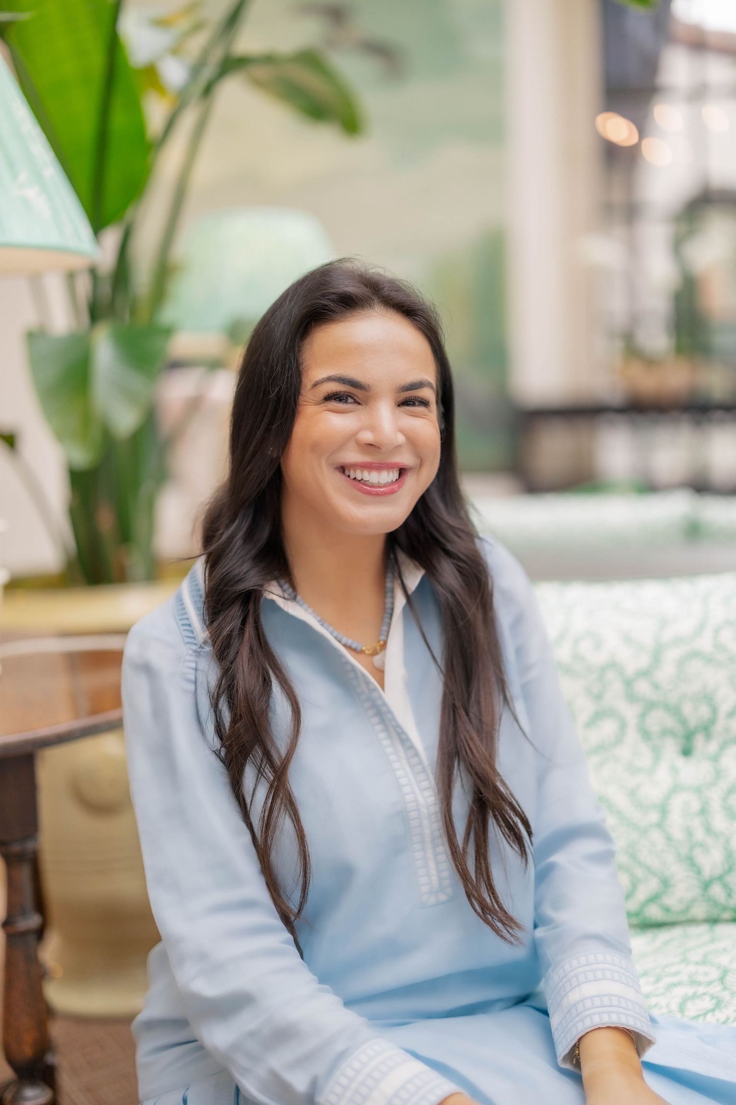 Smiling woman with long dark hair wearing a light blue linen outfit seated on green patterned sofa.