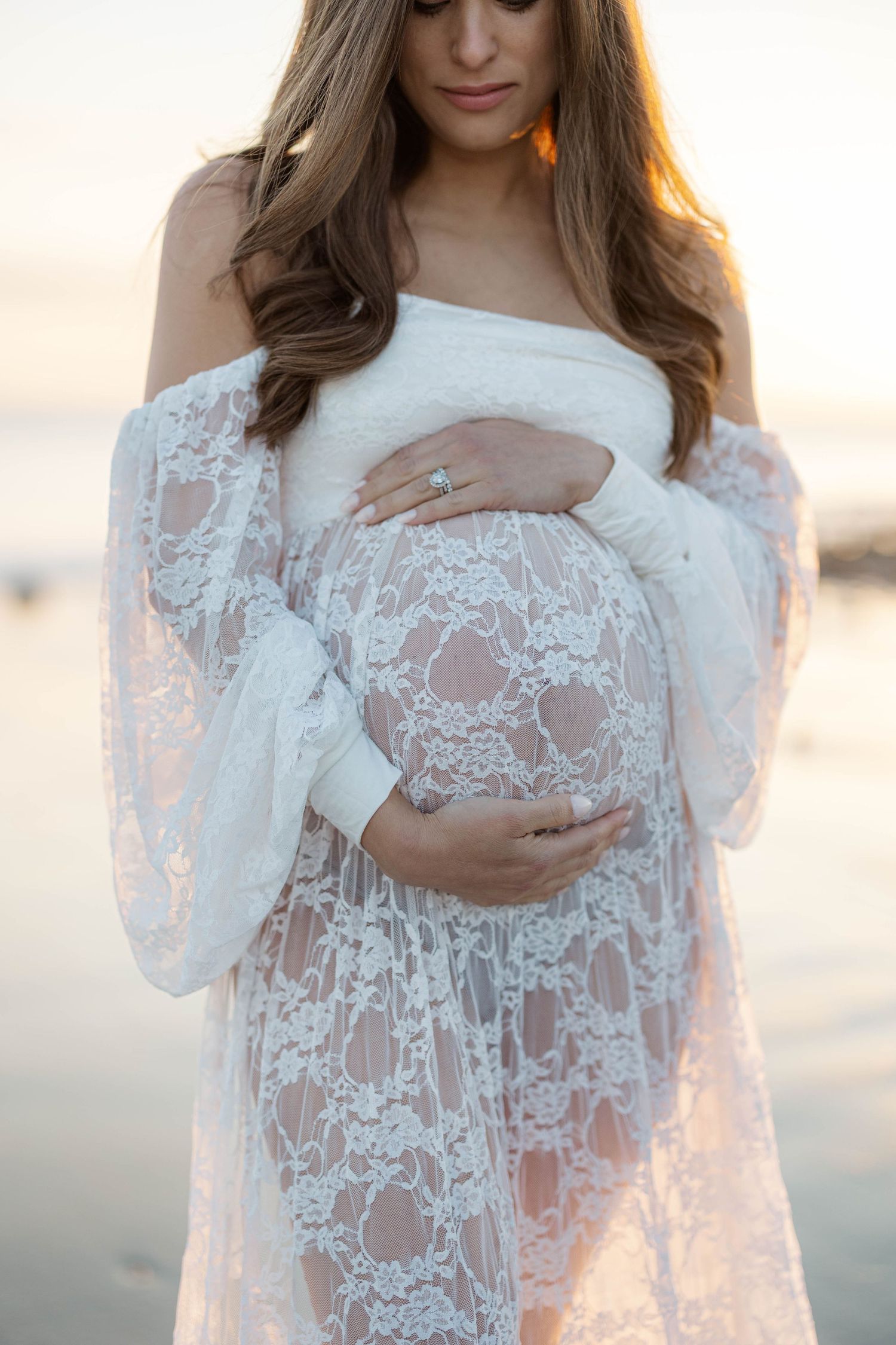 Pregnant woman in white lace off-shoulder gown cradling baby bump at sunset beach, maternity photography.