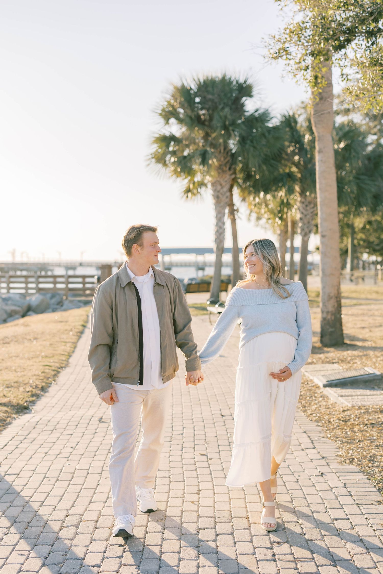 Pregnant couple holding hands walking on a brick path surrounded by palm trees in golden hour sunlight.