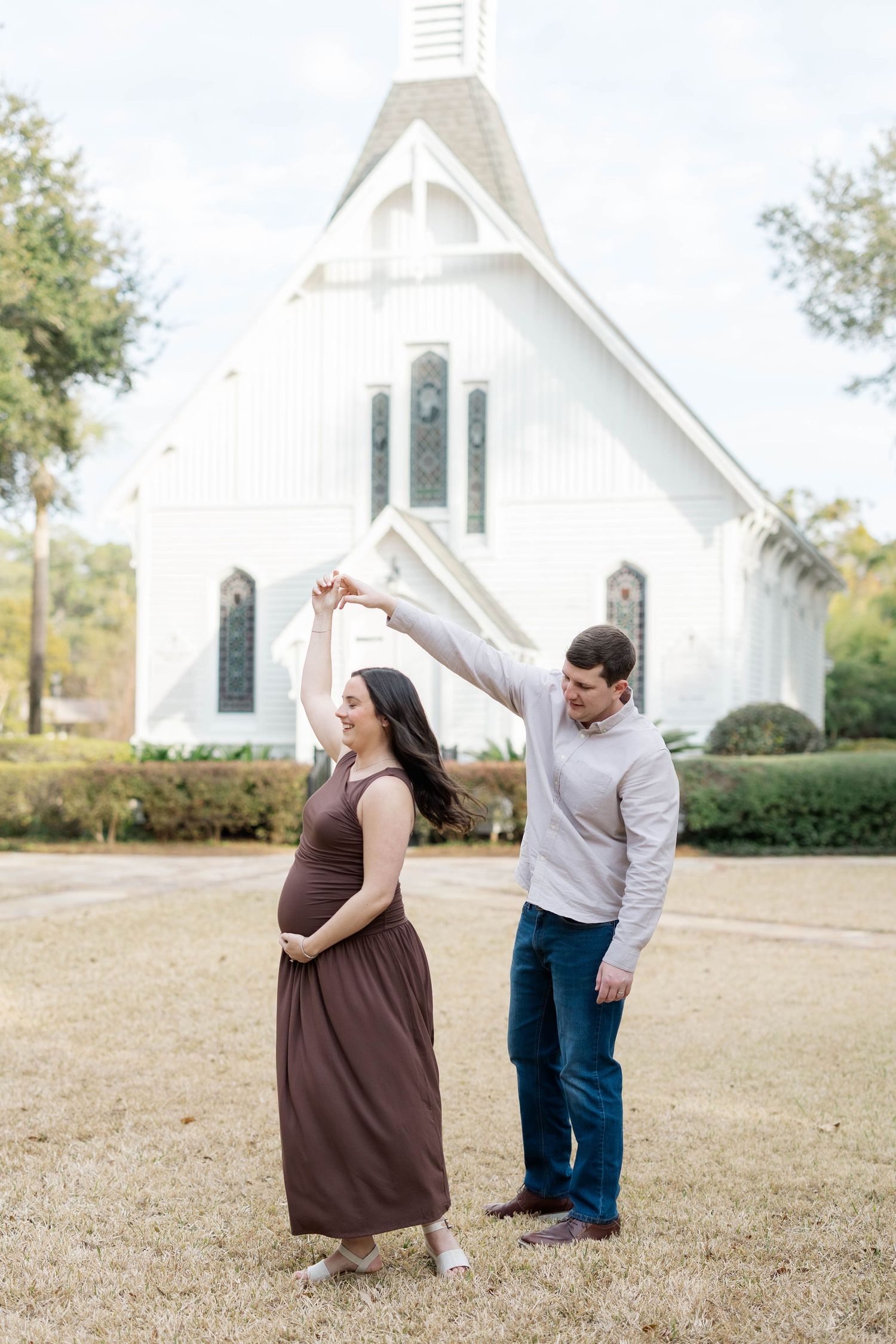 Pregnant couple dancing outdoors in front of a white chapel, woman in brown maxi dress cradling baby bump.