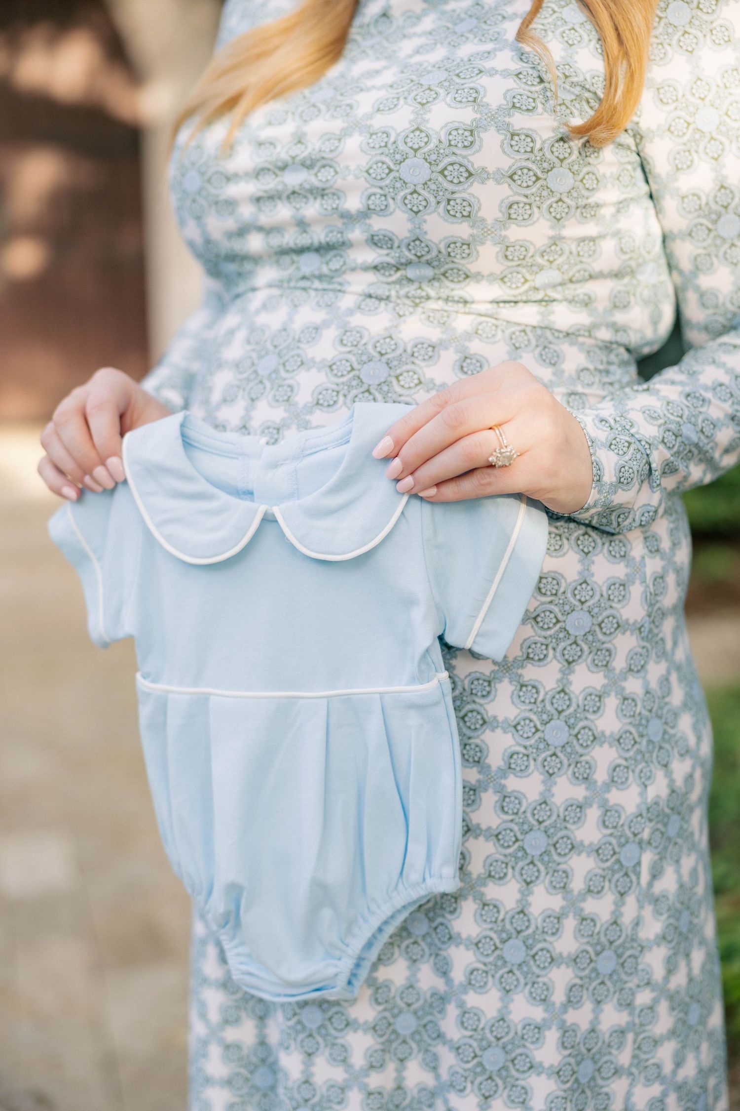 Pregnant woman in floral dress holding a light blue baby boy bubble romper with peter pan collar outdoors.