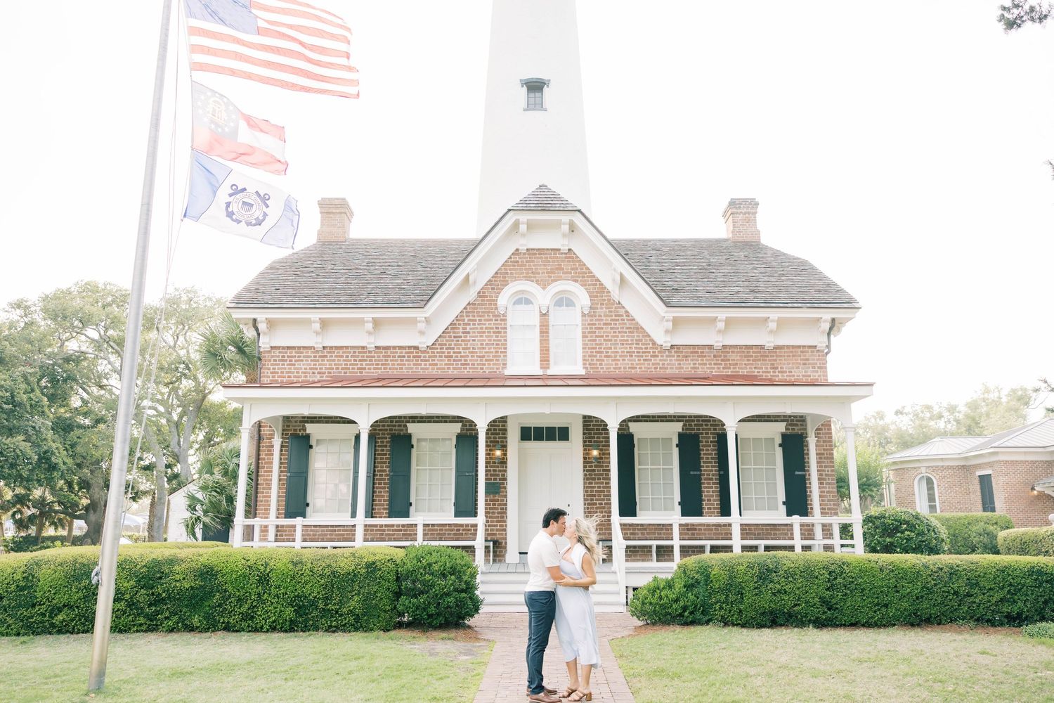 Couple embracing in front of a historic brick lighthouse keeper's house with American flag, lush greenery, and white lighthouse tower.