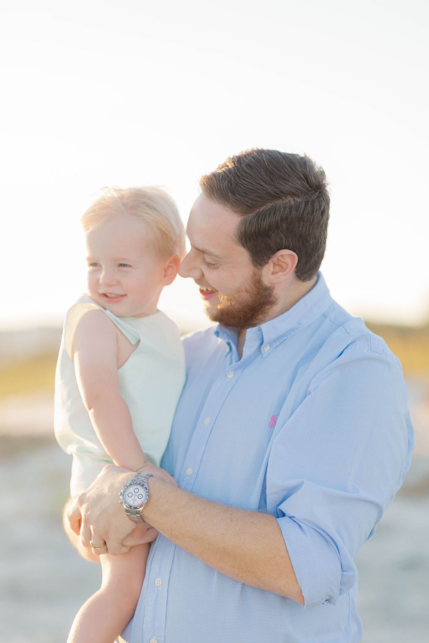 A bearded father in a blue shirt tenderly holds his smiling toddler on a sunny beach.