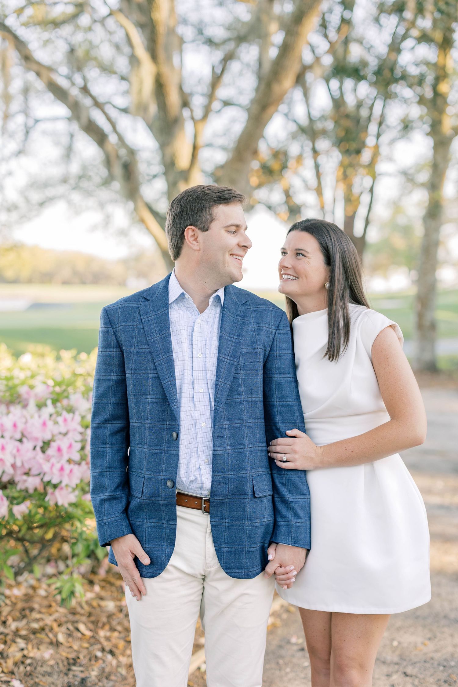 Couple smiling at each other outdoors, man in blue blazer and woman in white dress, with pink flowers and trees.