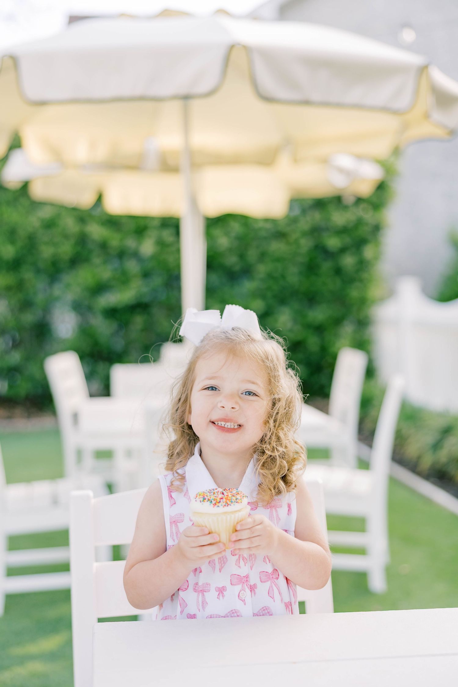 Young girl in pink flamingo dress holding colorful sprinkle ice cream cup outdoors under white patio umbrella.