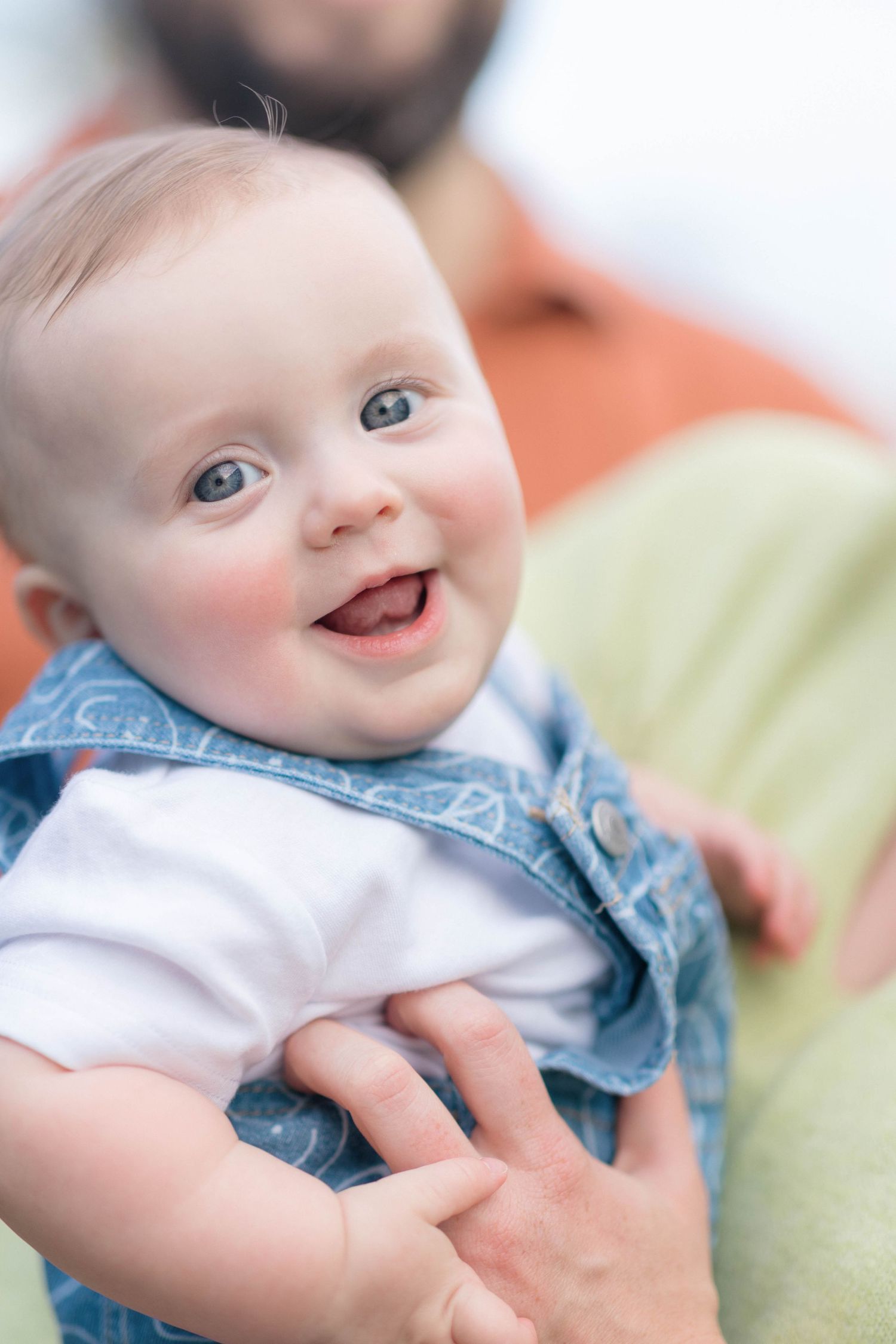 Happy smiling baby with blue eyes wearing a denim vest, being held by an adult in a warm, bright setting.