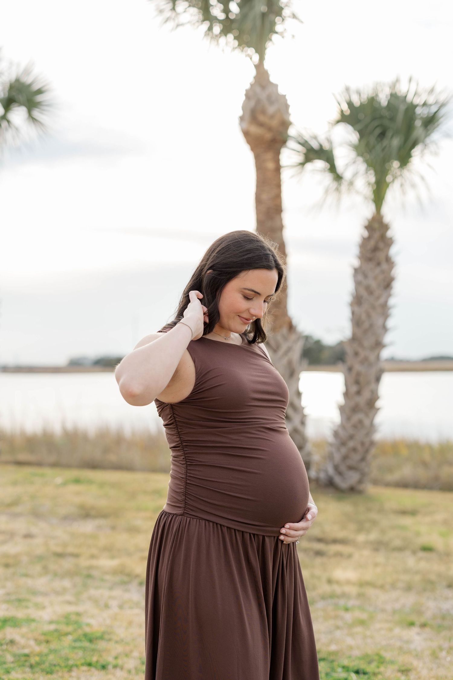 Pregnant woman in brown dress cradling baby bump near palm trees by water.