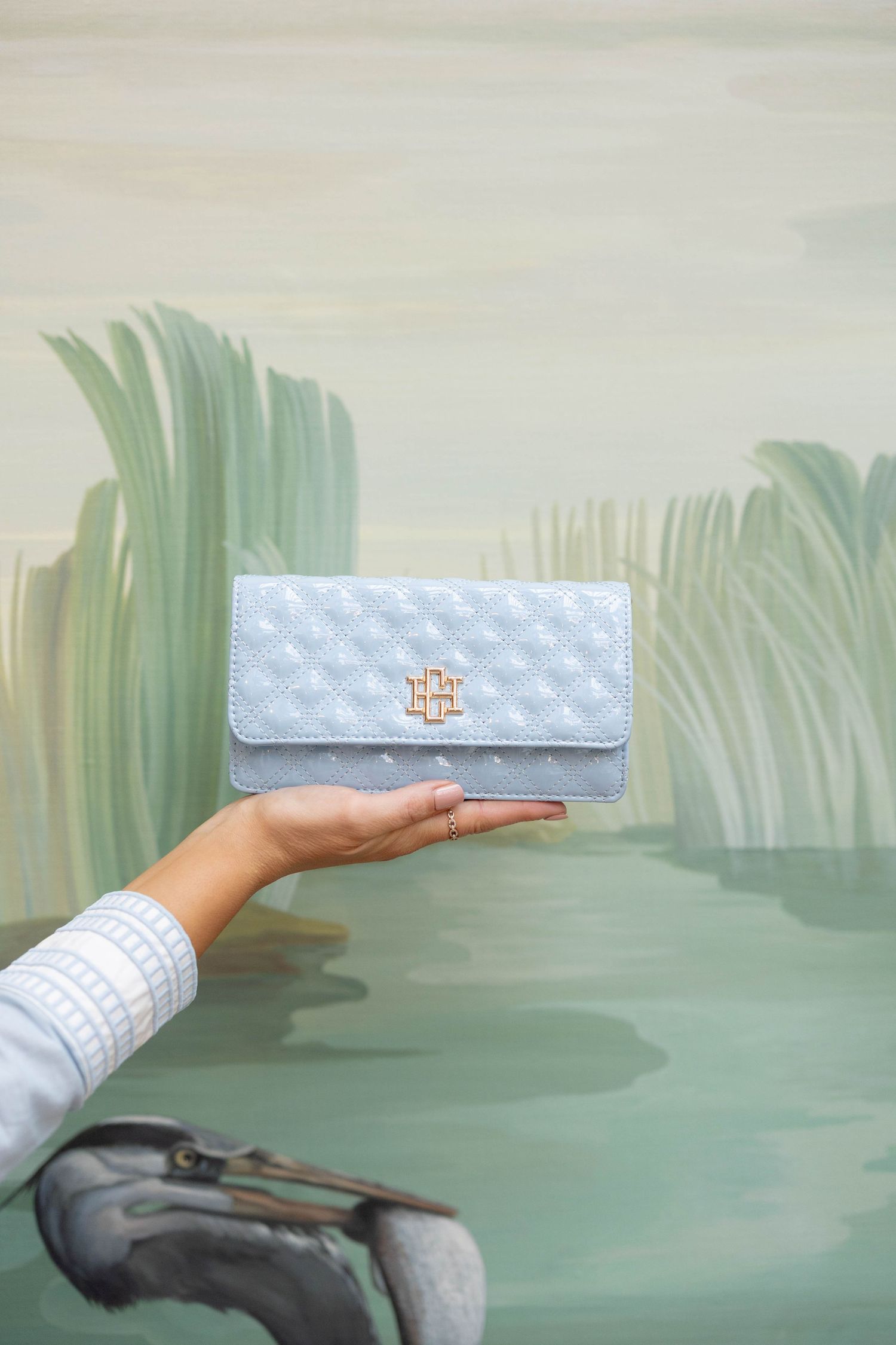 Light blue quilted wallet with gold logo held by hand against a painted mural background.