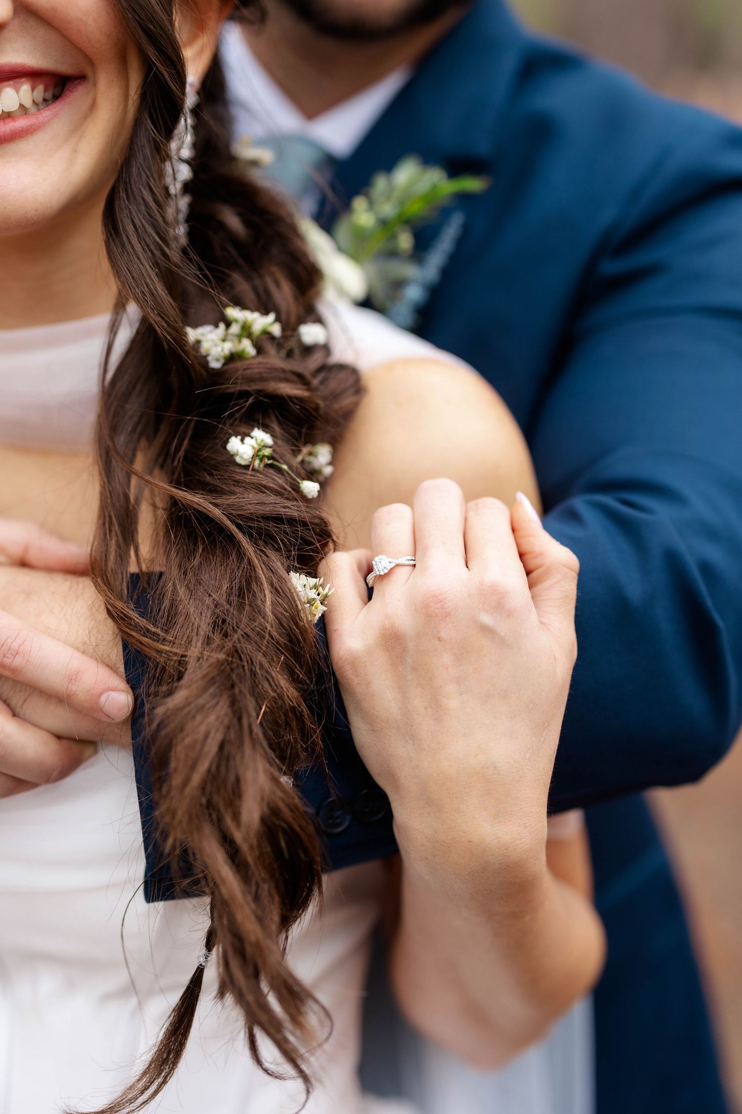 Close-up of engagement ring on hand with small white flowers and navy suit jacket in background.