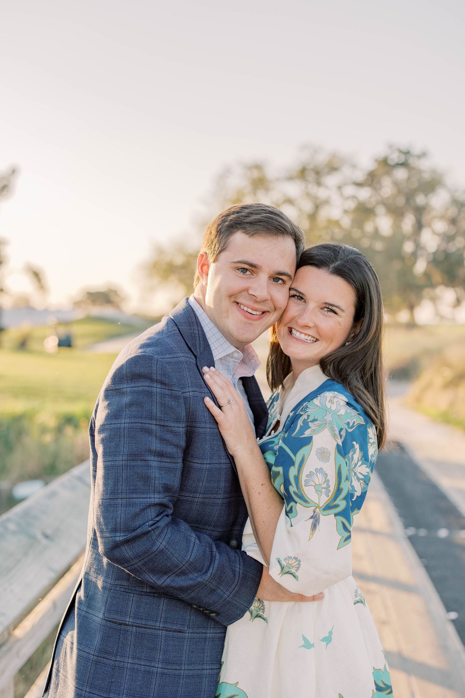 A smiling couple embraces on a wooden boardwalk at golden hour, man in blue plaid suit, woman in floral dress.