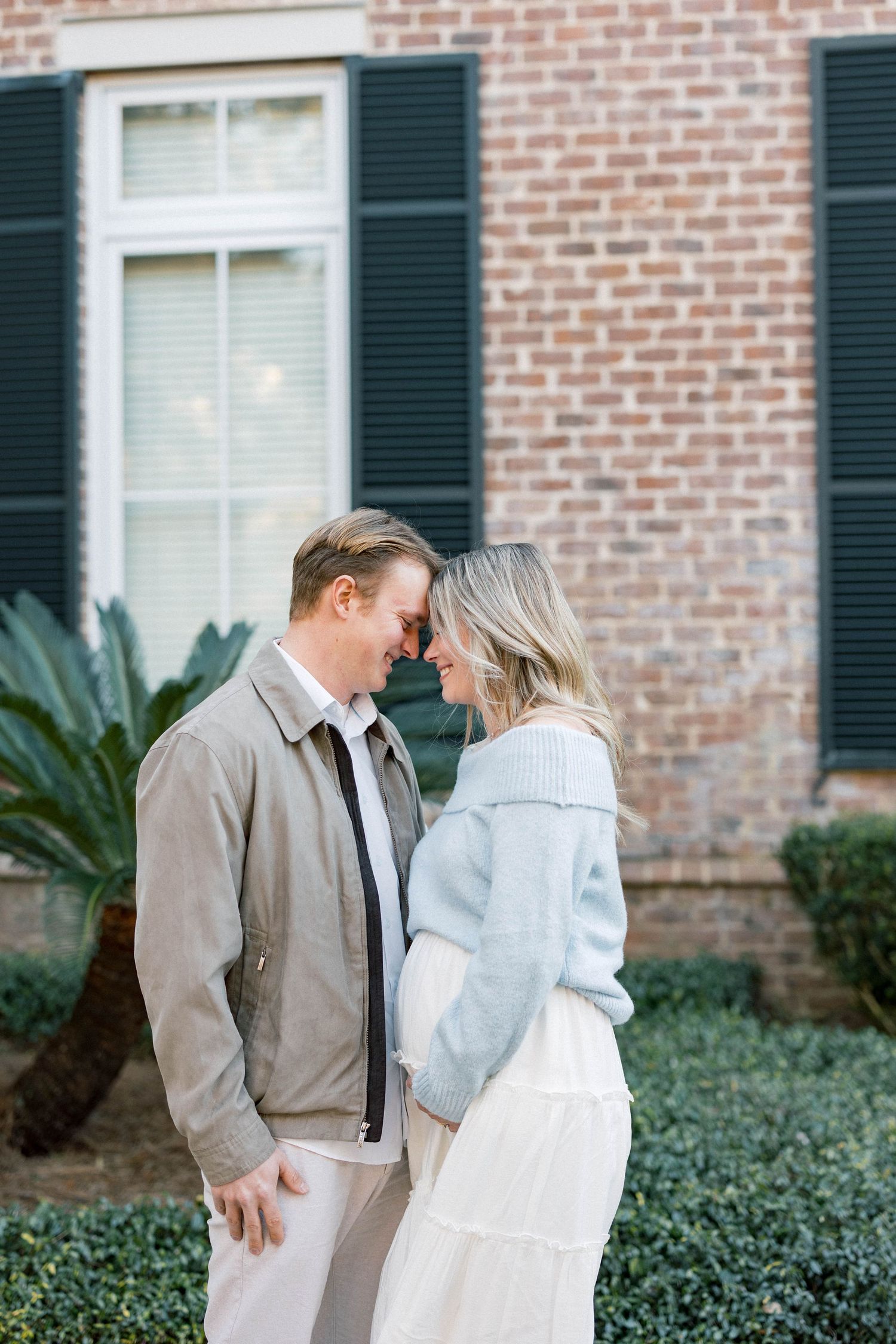 Expectant couple shares tender forehead-to-forehead moment outside brick home, woman in light blue sweater showing baby bump.