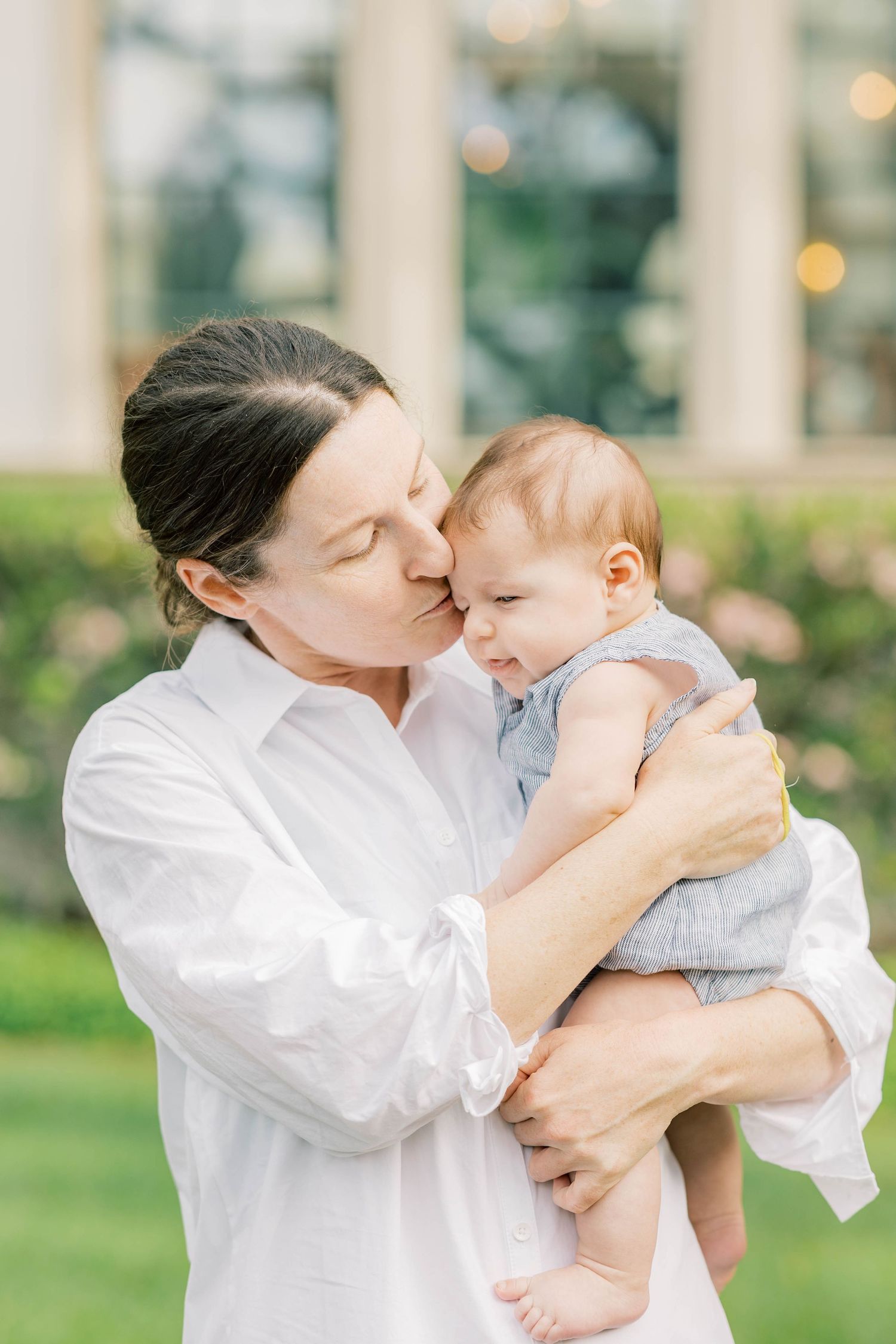 A woman in a white shirt tenderly kissing a baby outdoors near a garden.