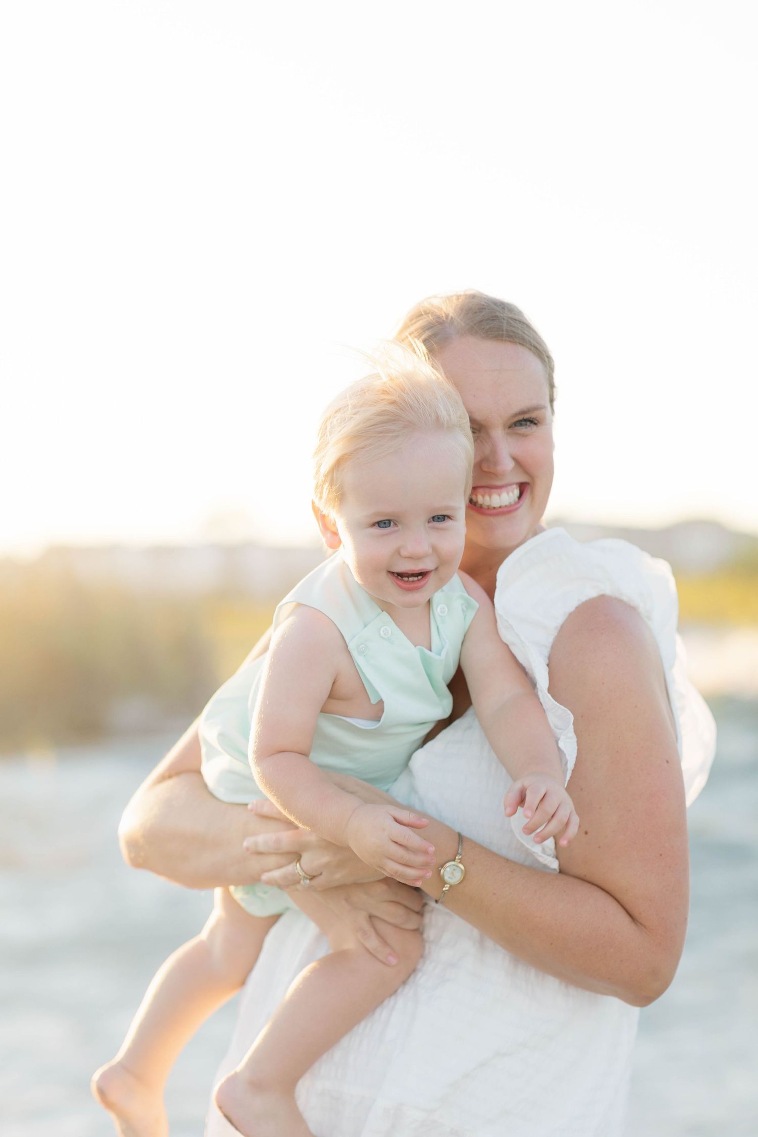 Smiling mother holding her happy baby girl on a sunny beach, both dressed in light pastel outfits.