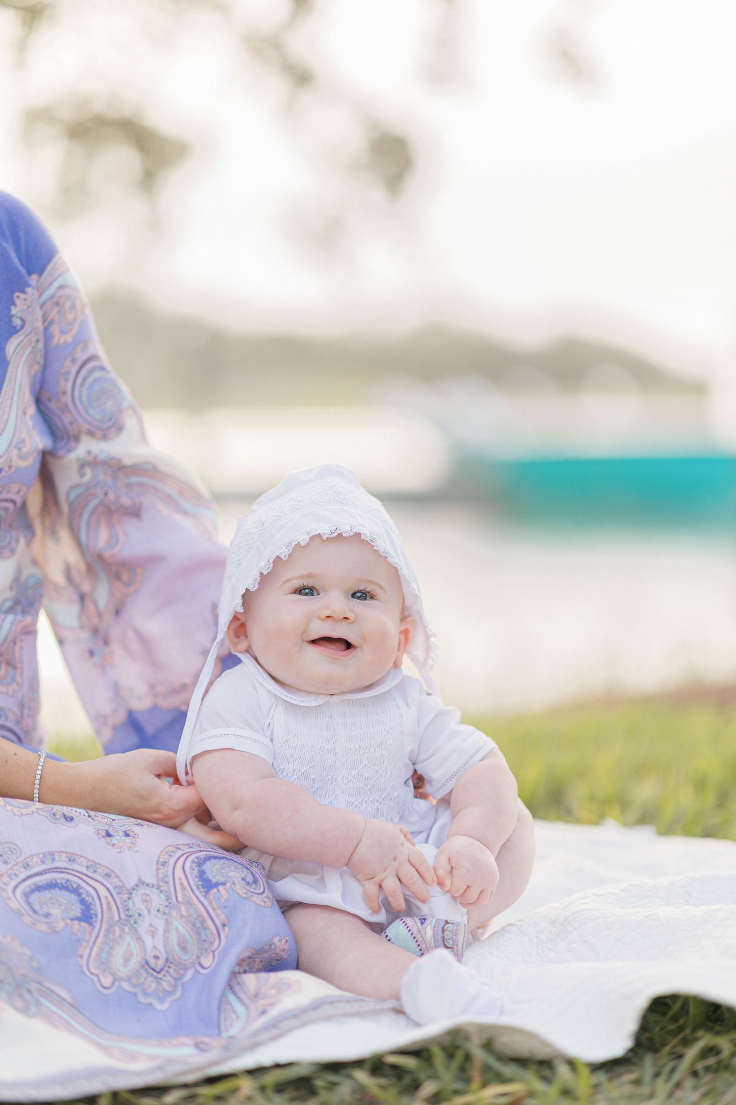 Smiling baby in white bonnet and outfit sitting on blanket outdoors near water with adult in floral dress nearby.