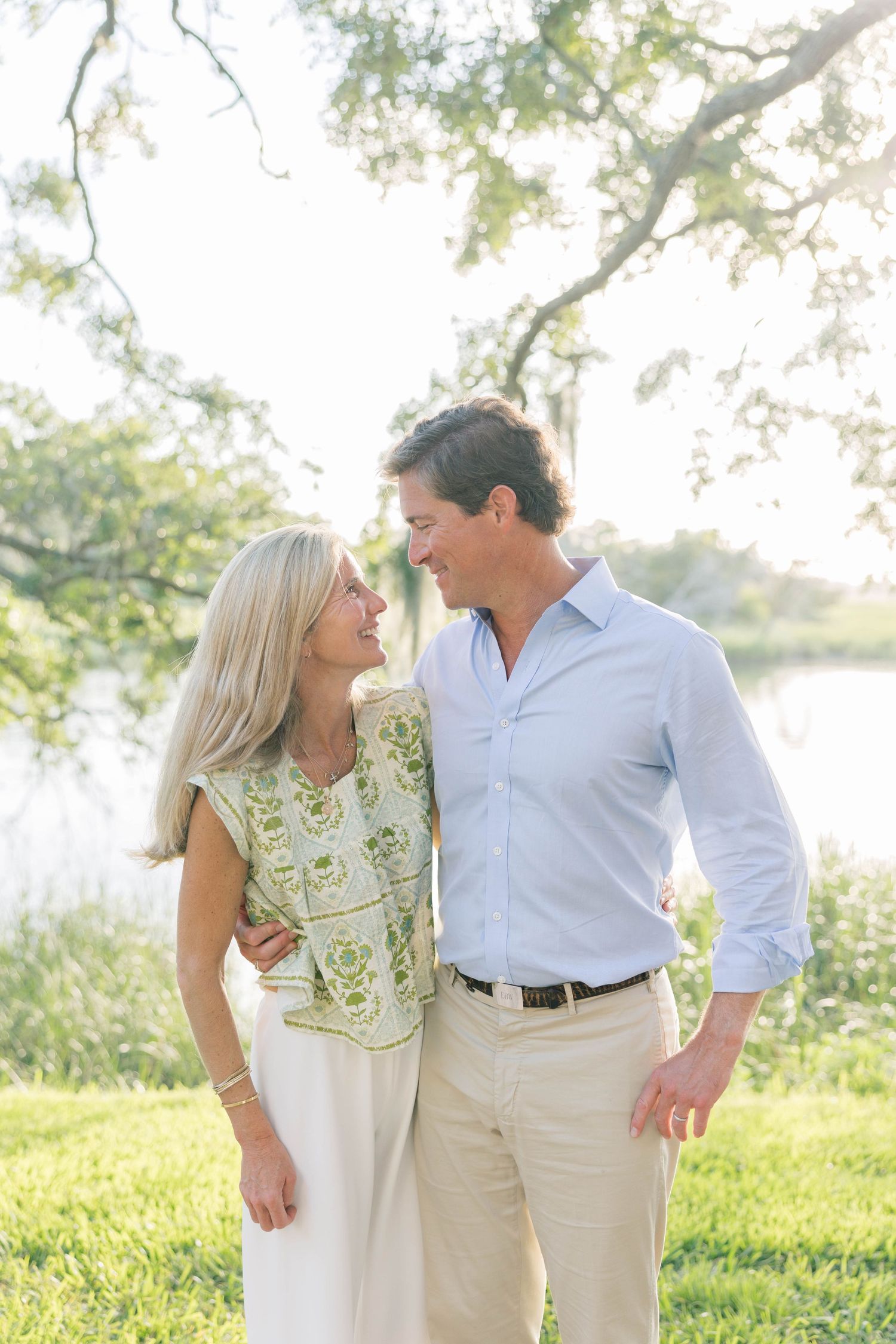 A smiling middle-aged couple embracing outdoors near a lake, surrounded by lush green trees in warm sunlight.