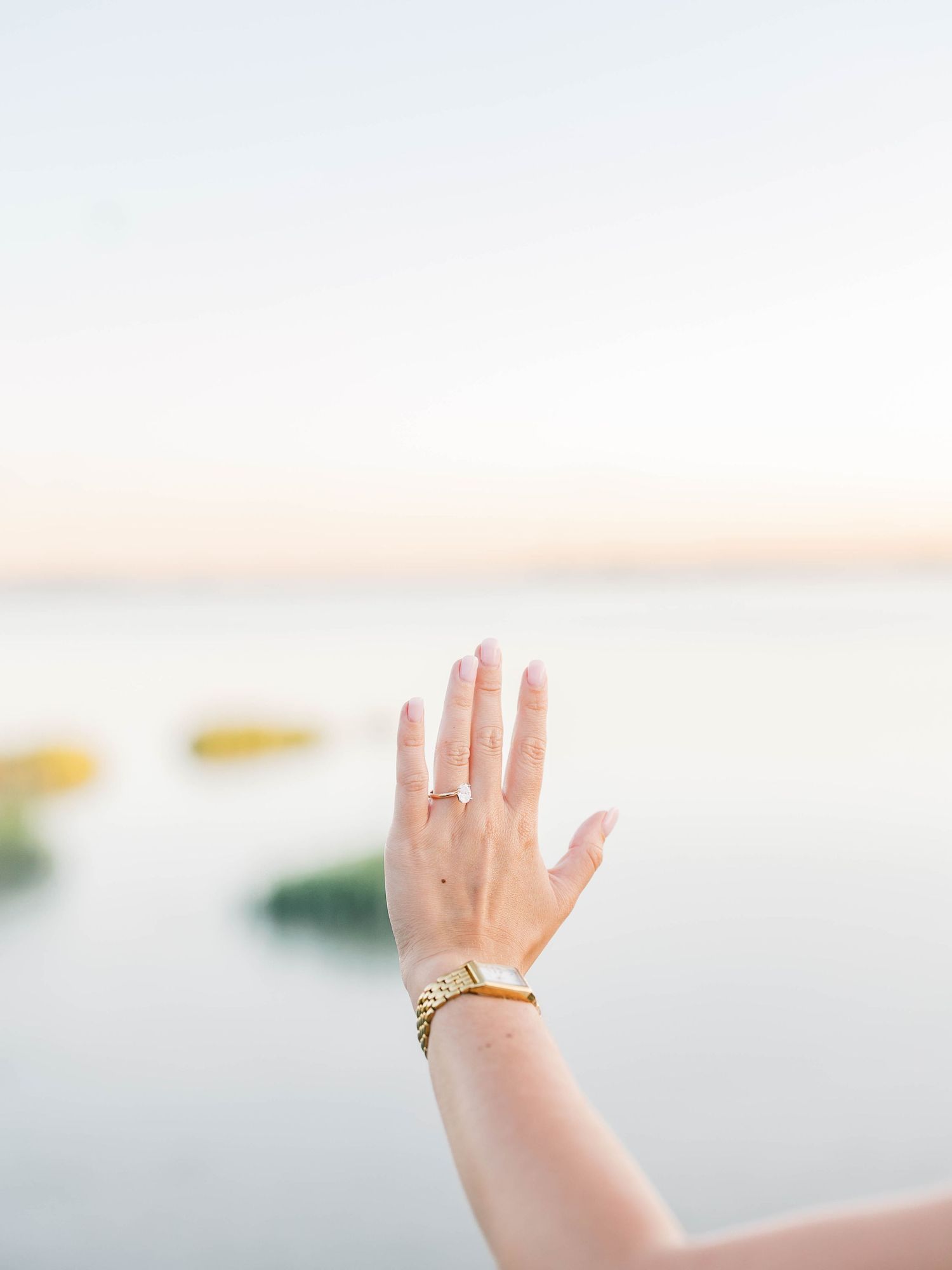 A woman's hand raised against a serene ocean sunset, wearing a gold watch and delicate ring.