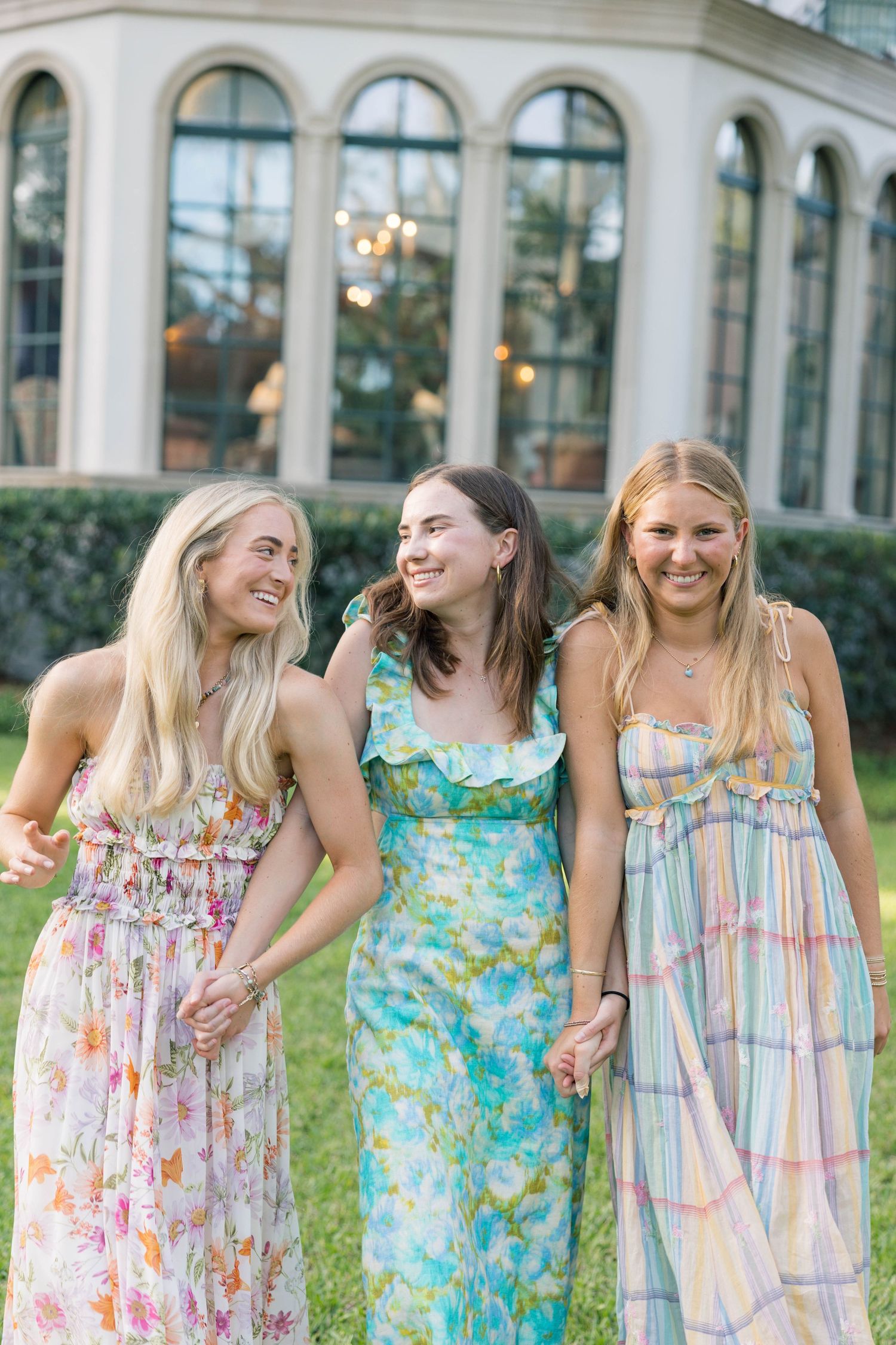 Three young women in colorful floral maxi dresses smiling and holding hands outdoors near an elegant building.