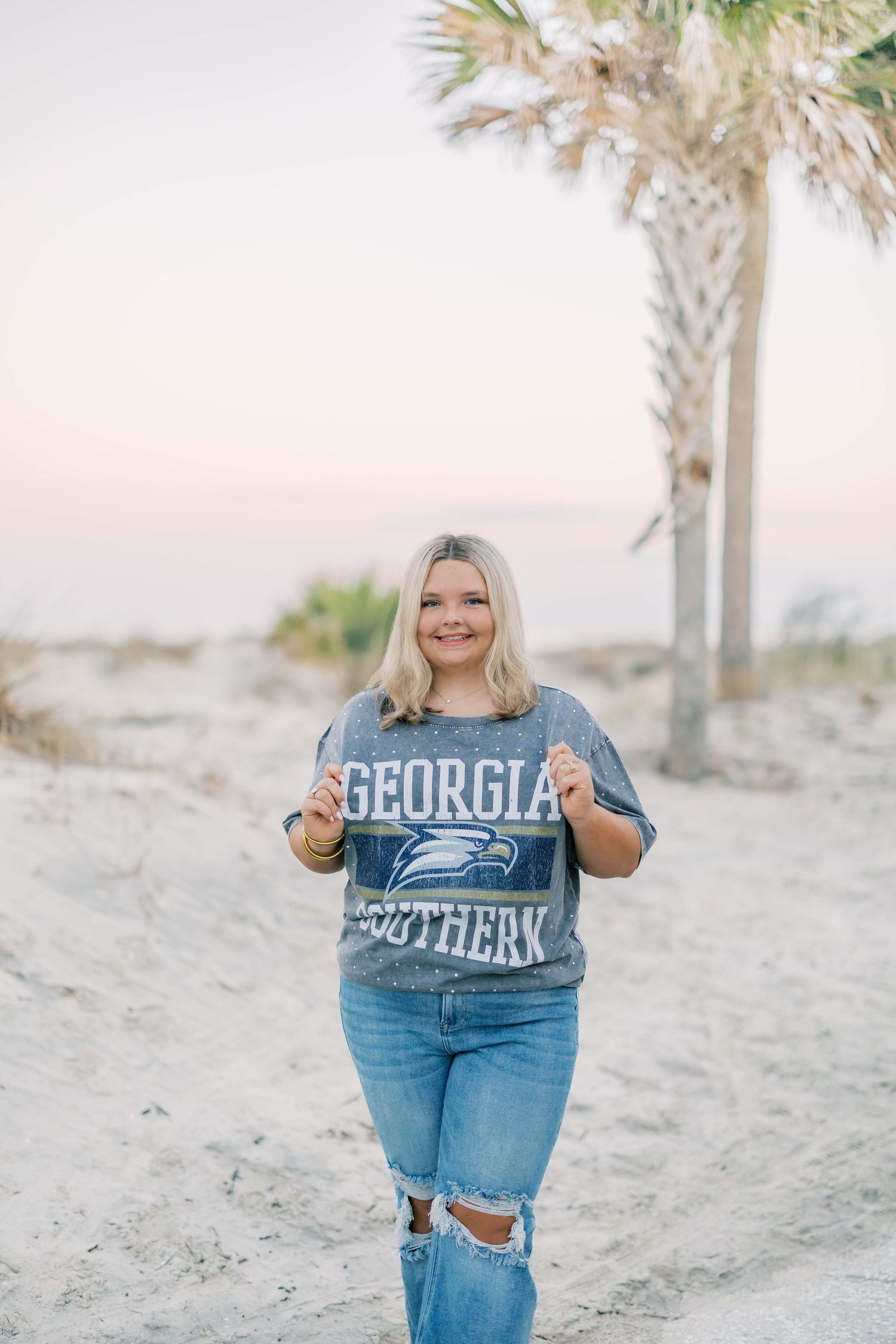 Smiling blonde woman wearing a Georgia Southern Eagles sweatshirt and ripped jeans on a sandy beach near palm trees.