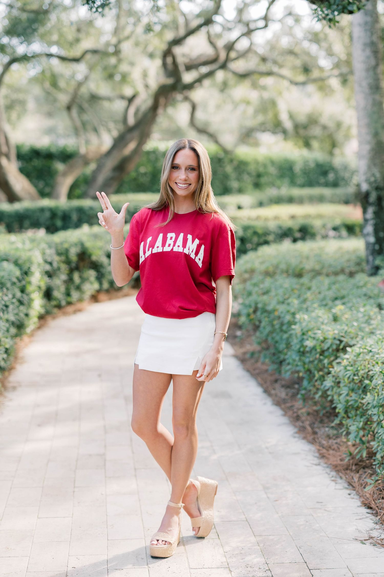 Woman in red Alabama t-shirt, white mini skirt, and wedge sandals posing on a tree-lined brick pathway.