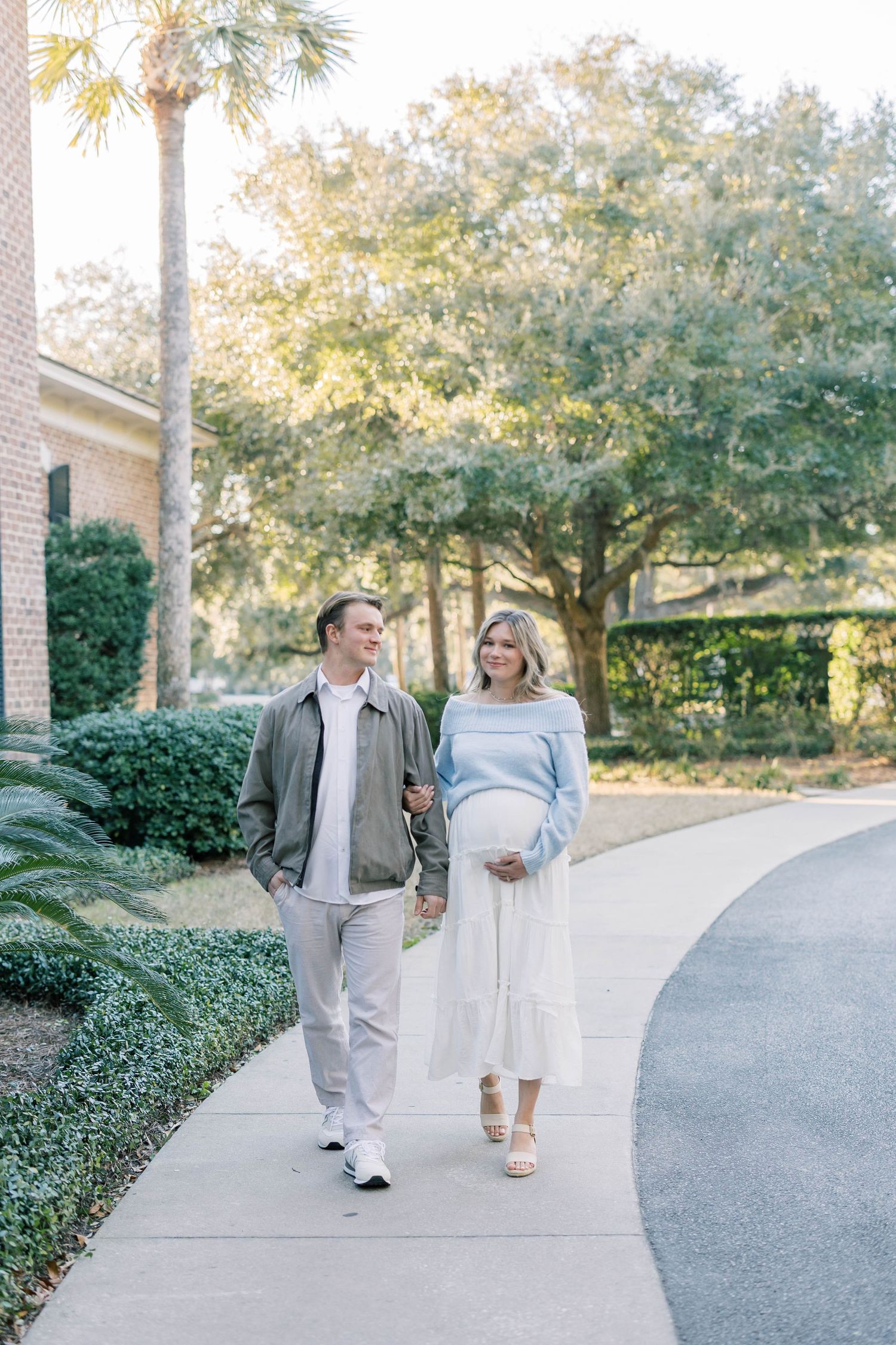 Couple walking on a curved path, woman in light blue sweater and white skirt showing baby bump, man in gray jacket.