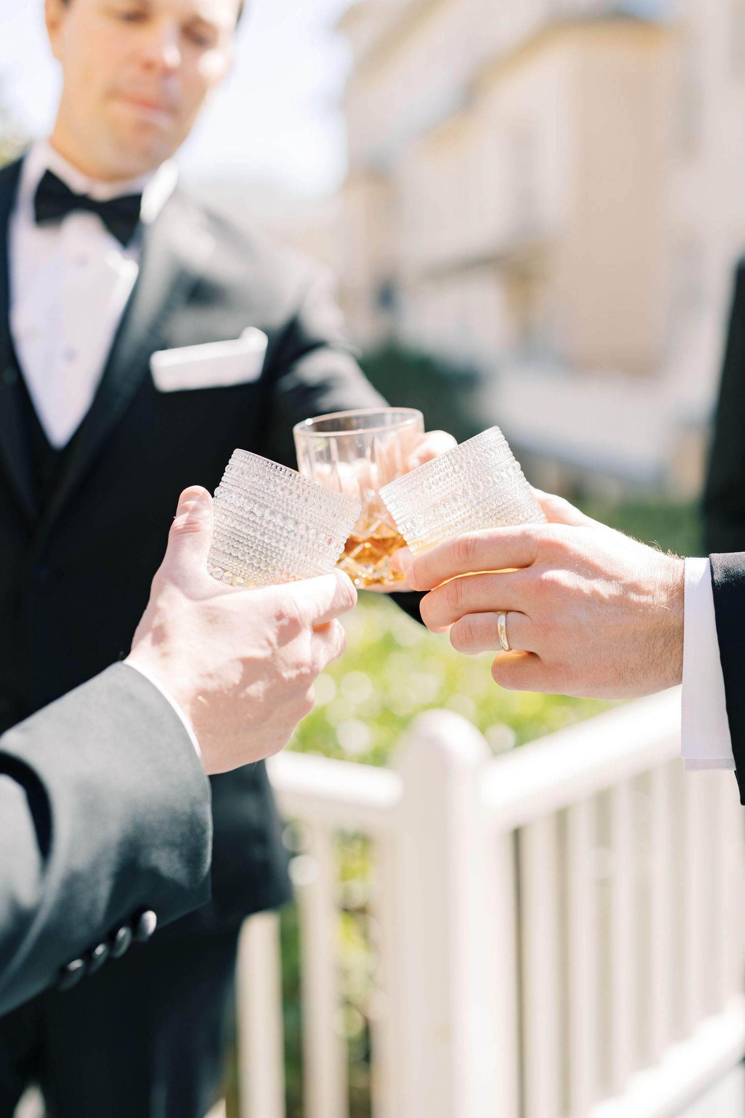 Men in tuxedos toasting with crystal glasses outdoors at a wedding celebration in bright sunlight.