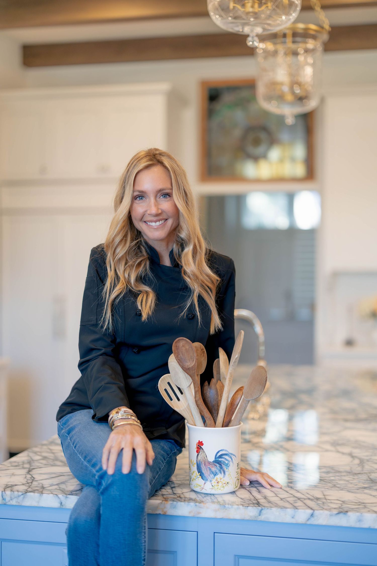 Smiling blonde woman sitting on a marble kitchen island beside a rooster utensil holder with wooden spoons.