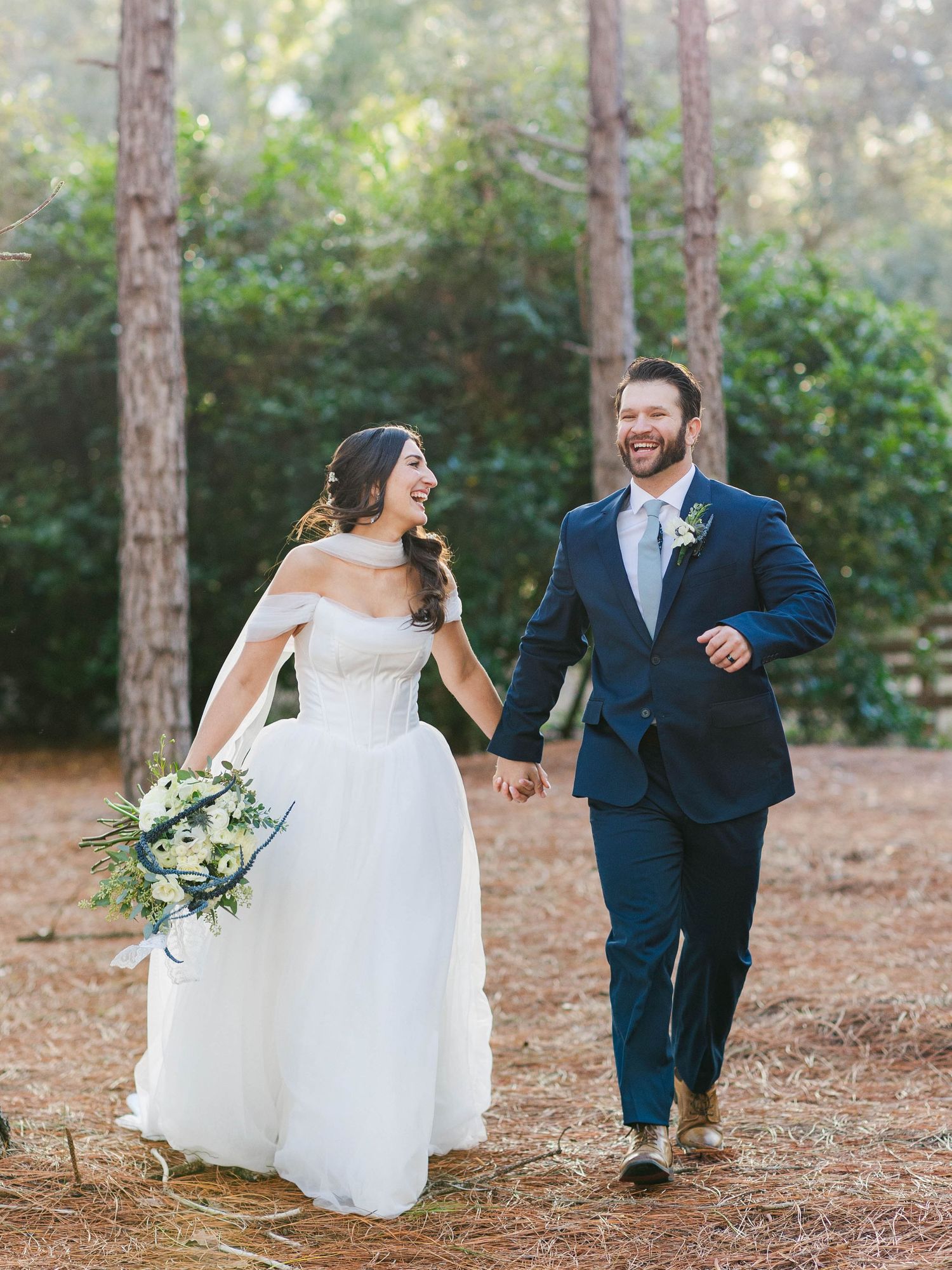 Couple in wedding attire playfully runs through a pine forest while holding hands and laughing.