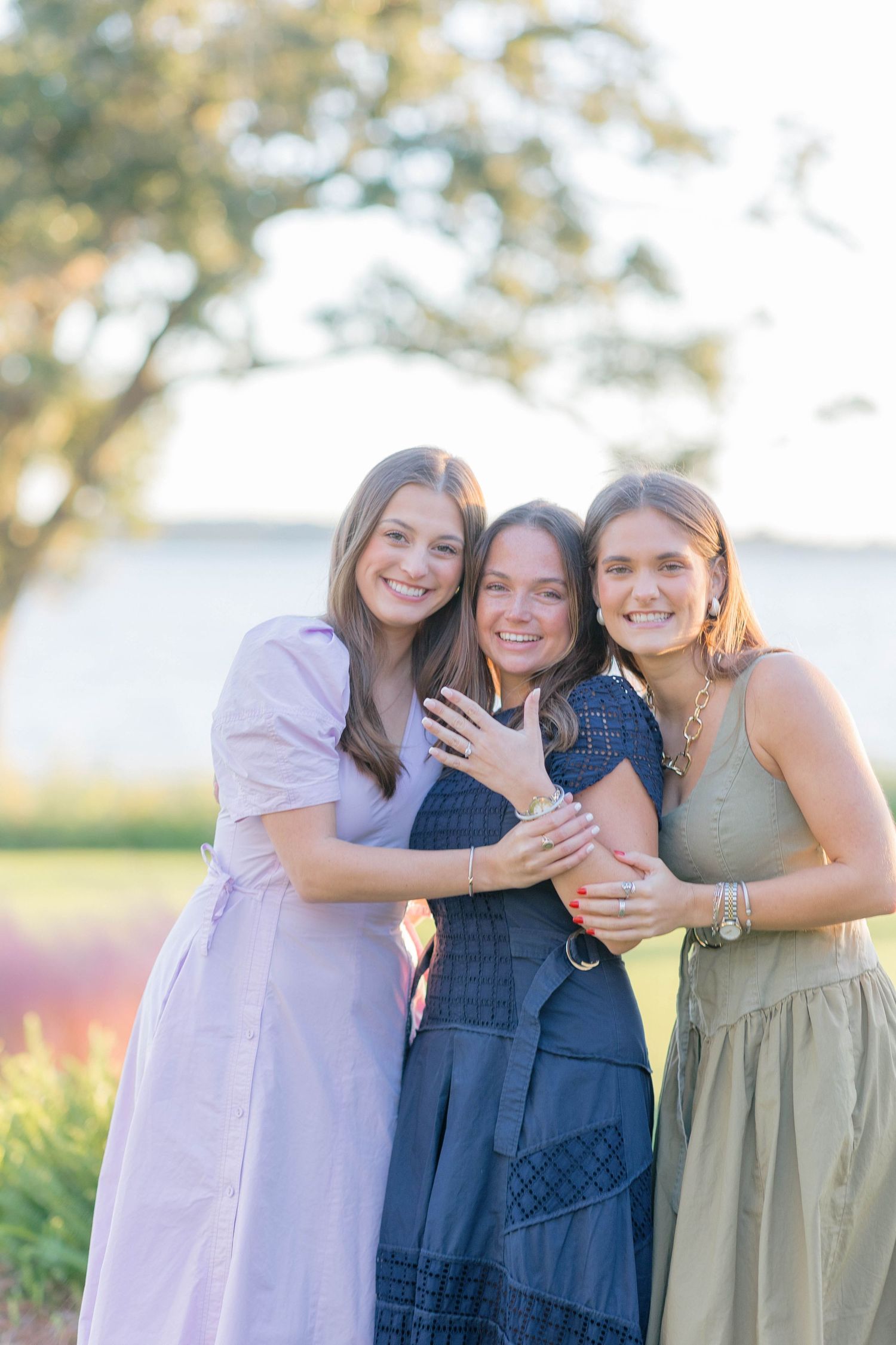 Three women hugging and smiling outdoors, one showing off engagement ring near scenic waterfront.