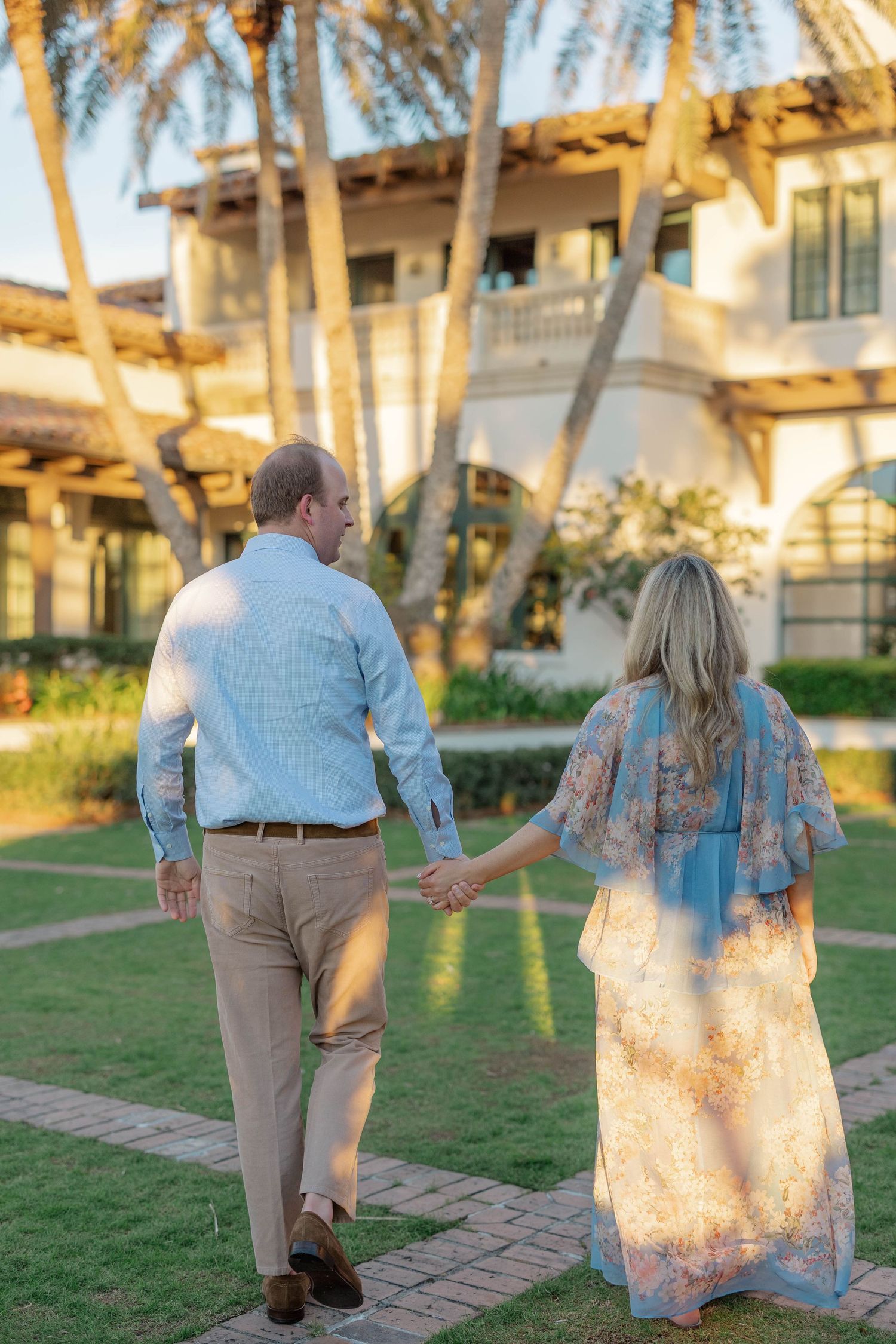 Couple holding hands walking toward a tropical villa surrounded by palm trees at golden hour.