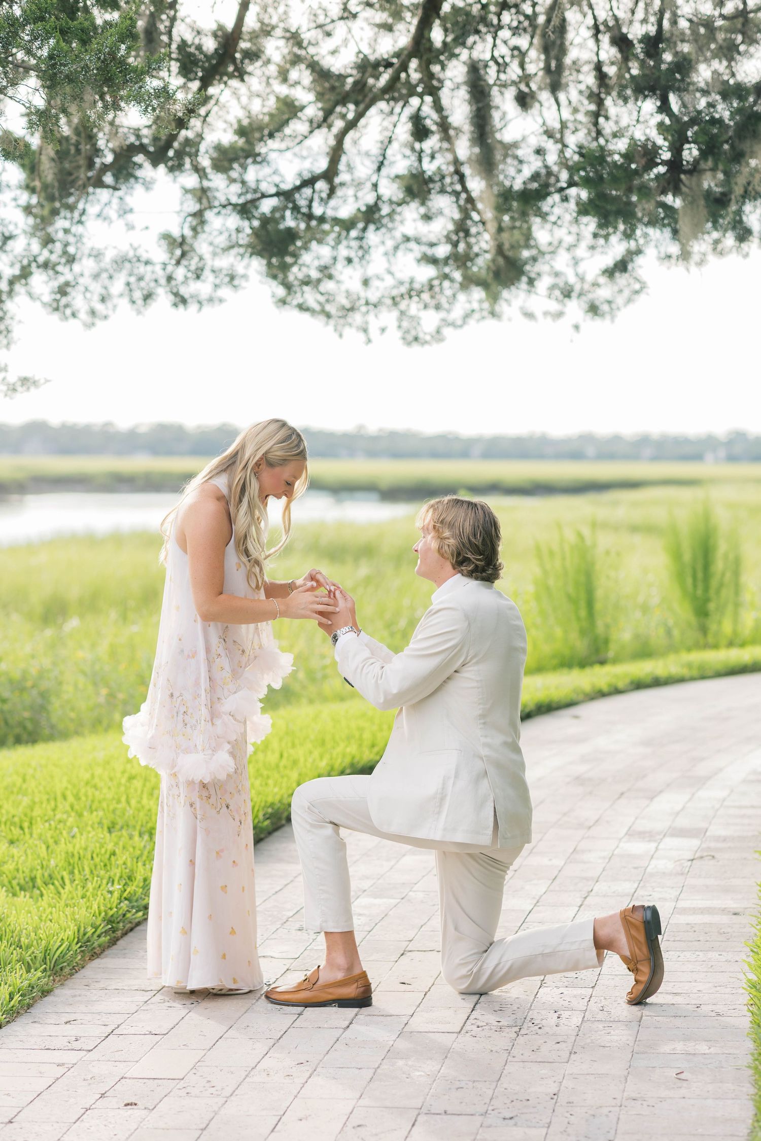 Romantic outdoor marriage proposal with man on one knee placing ring on womans finger near tranquil marsh.